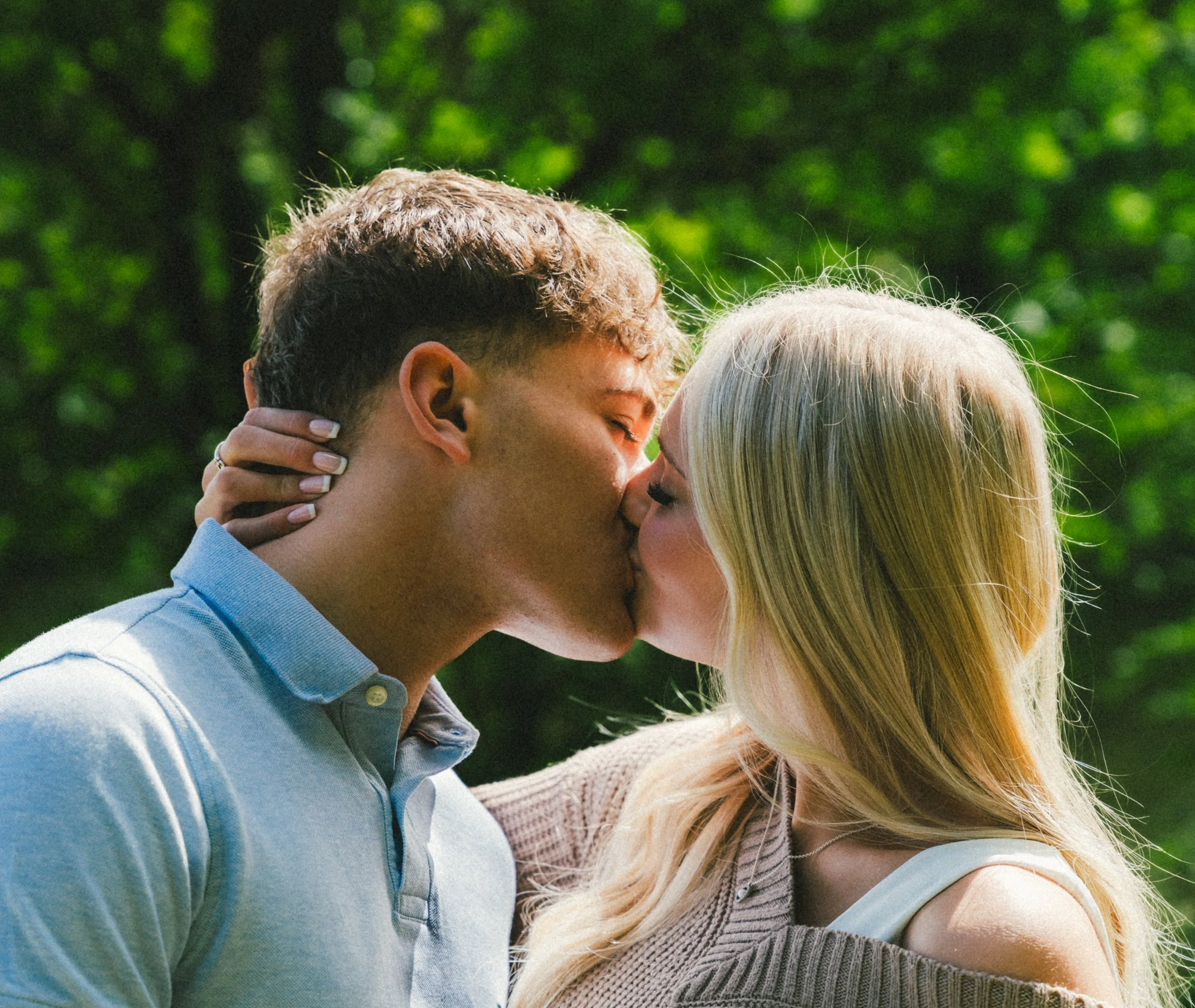 A young man and woman sharing a kiss outdoors, with green trees in the background.