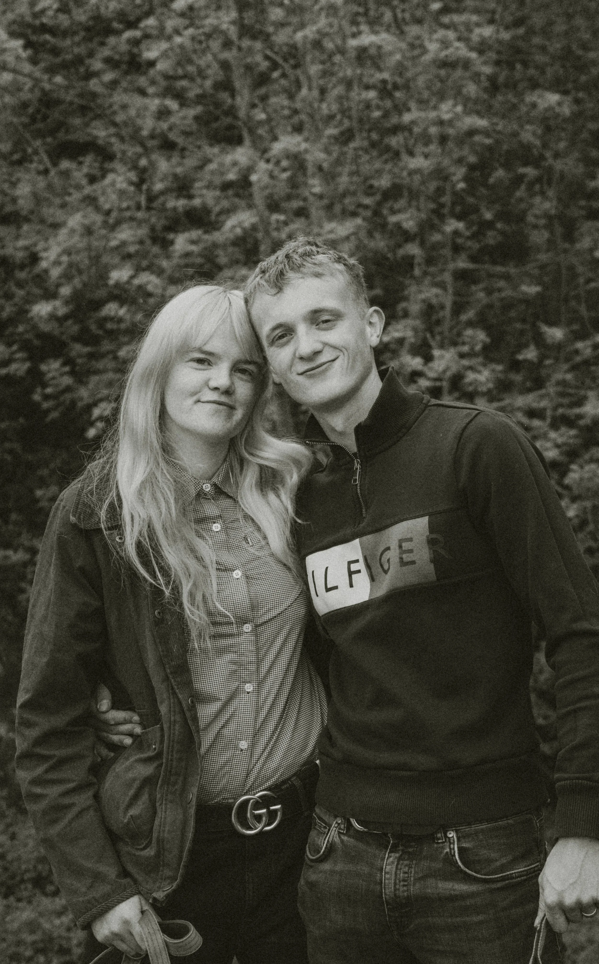 A black and white photo of a young man and woman smiling and standing close together in an outdoor setting with trees in the background. The woman has long blonde hair and is wearing a denim jacket over a checkered shirt. The man has short curly hair