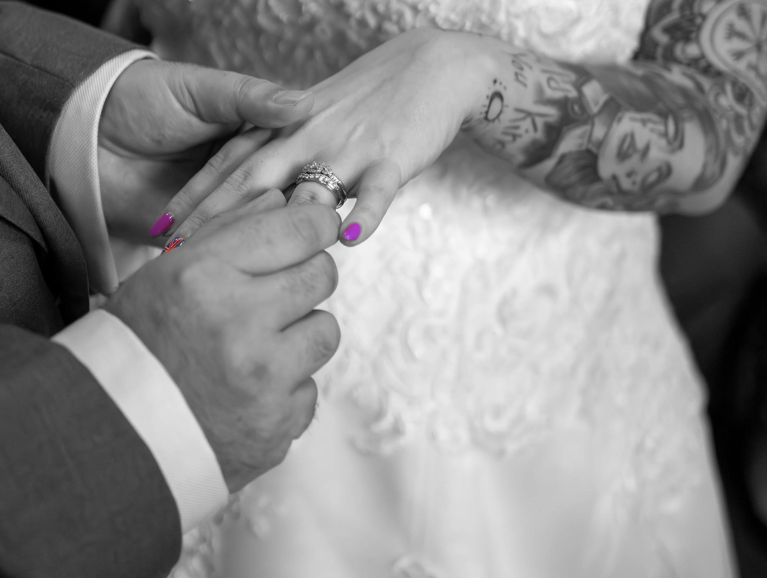 Black and white photo of a couple's hands holding each other during their wedding; the bride's hand shows a diamond engagement ring and a wedding band, with pink nail polish, and the groom's hand is holding her hand. The groom is wearing a suit and t