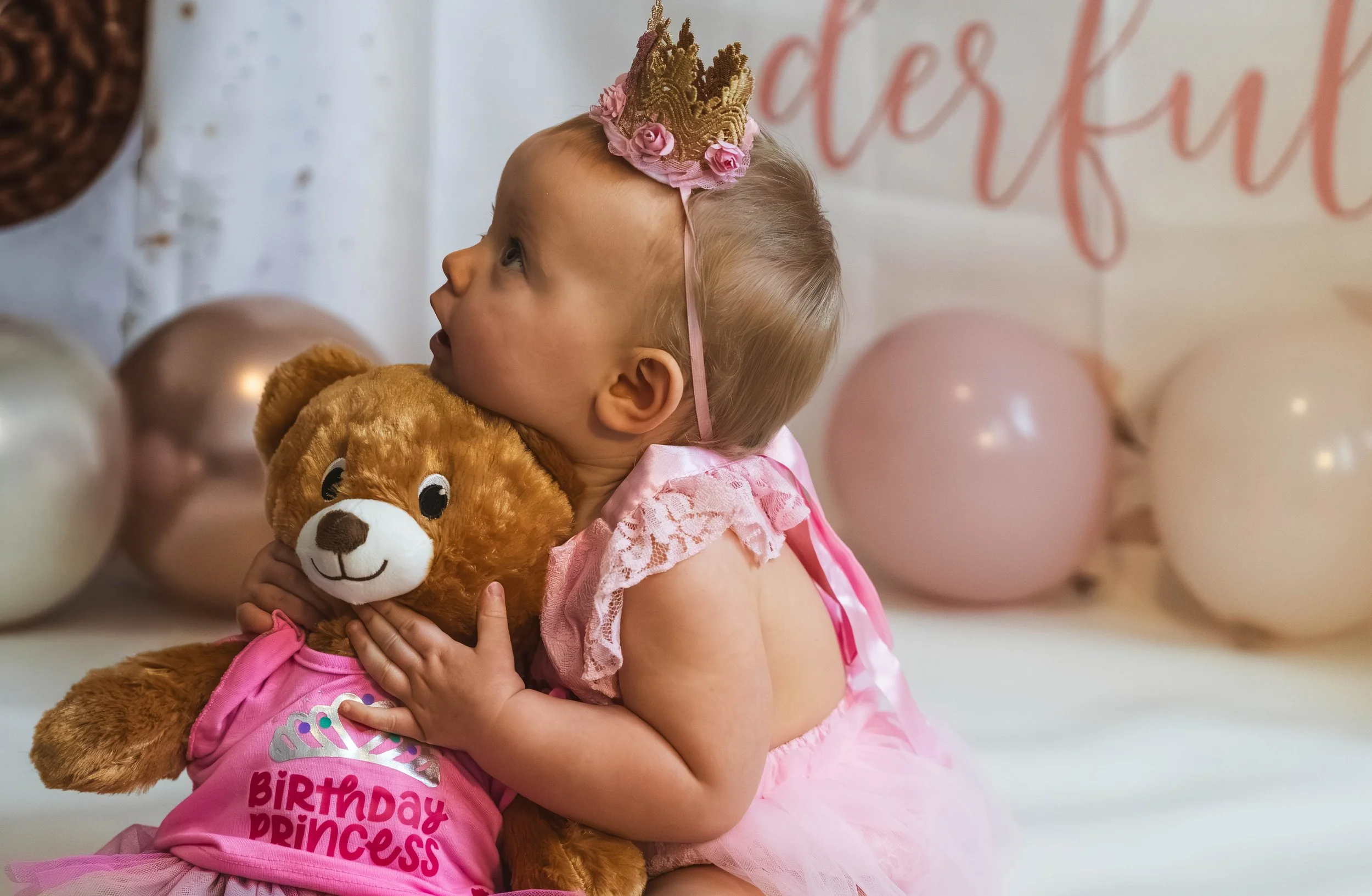 A young girl with blonde hair wearing a pink birthday dress and a crown, hugging a plush lion toy, at a birthday celebration with pink and white balloons and a sign in the background.