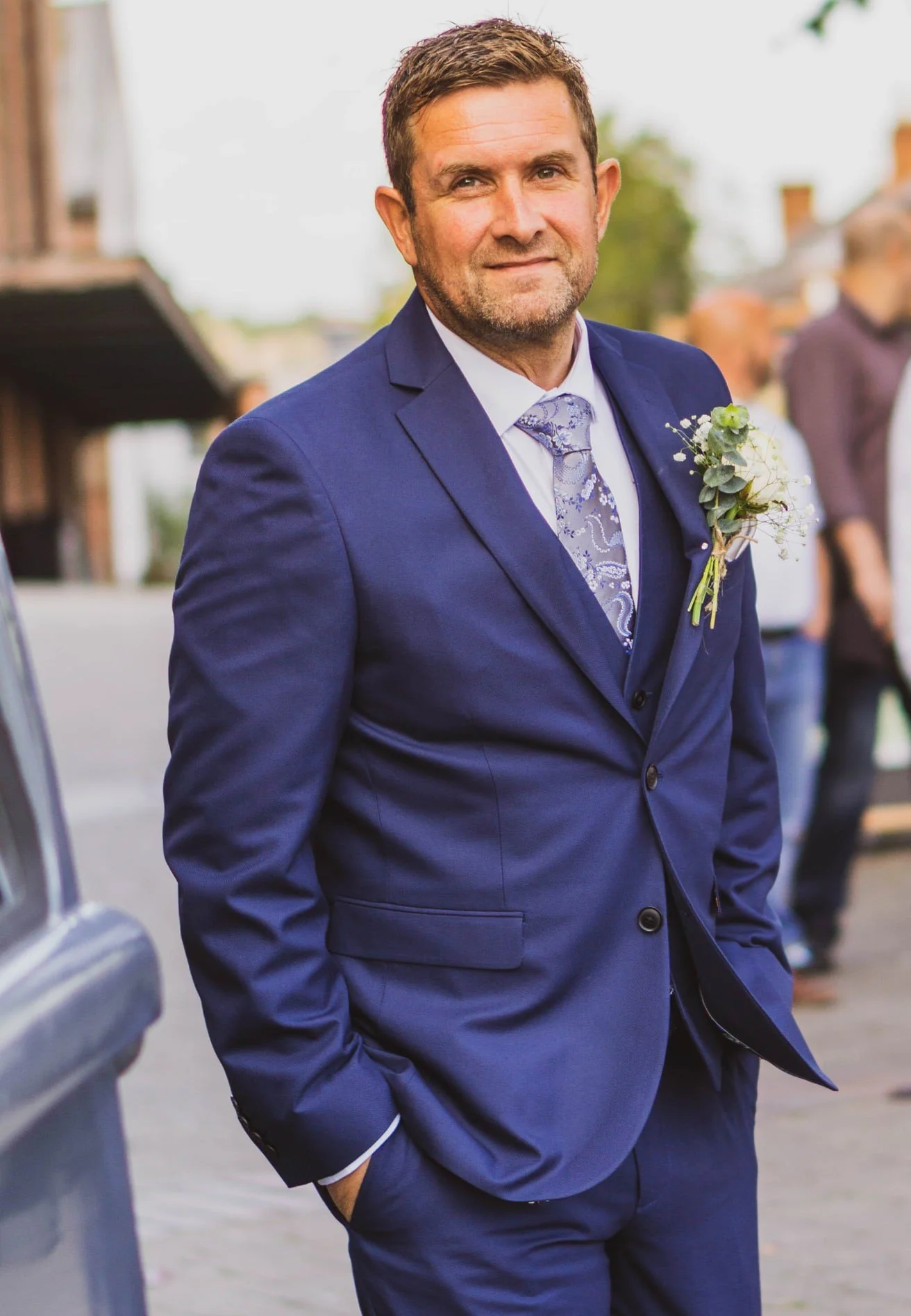 A man in a blue suit and purple tie standing outdoors with his hands in his pockets. He has short brown hair and a trimmed beard, and is wearing a boutonniere on his left lapel. In the background, there are blurred people and buildings.