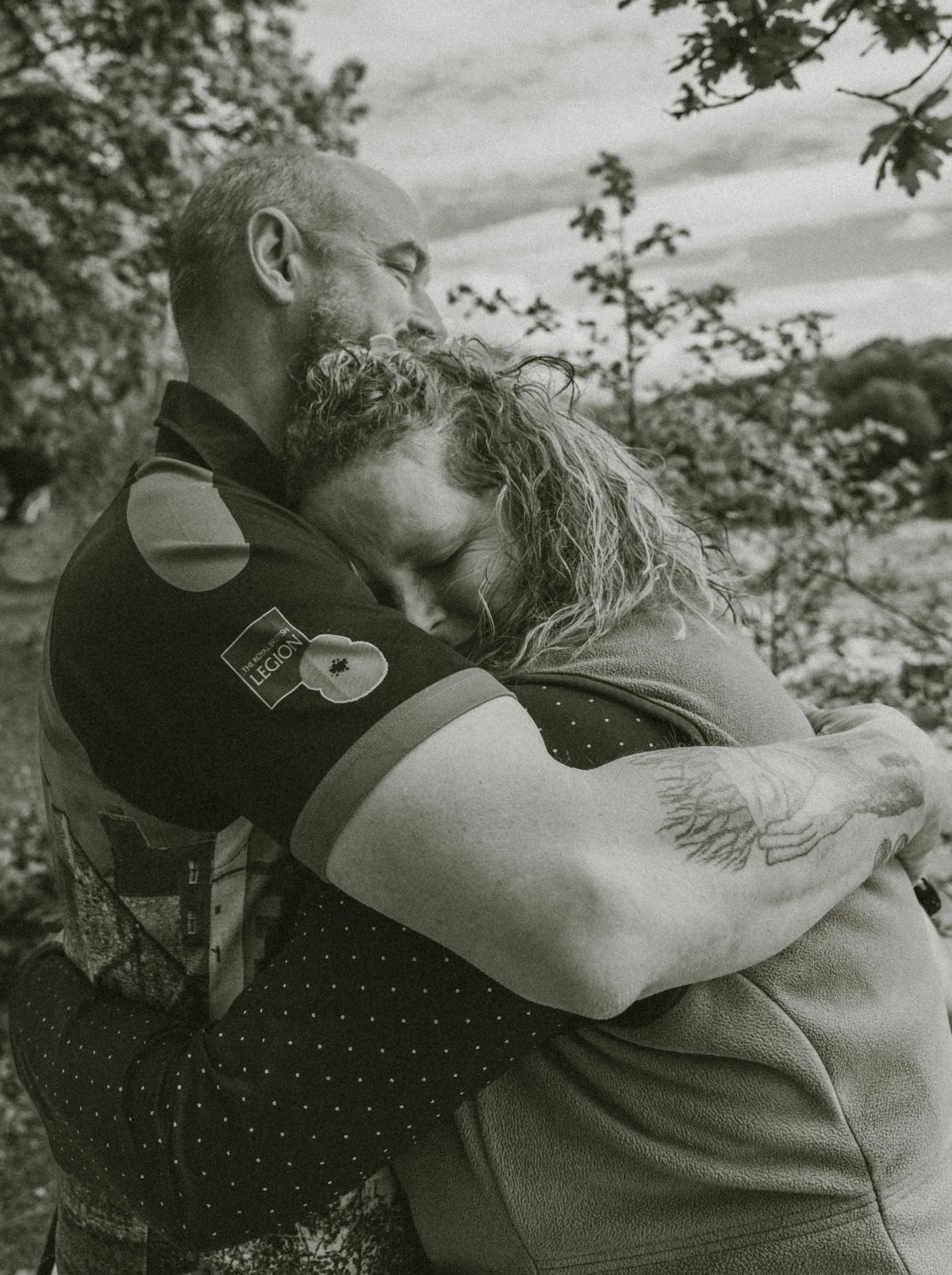 A man with a gray beard and short hair hugging a woman with curly hair outdoors, with trees and cloudy sky in the background.