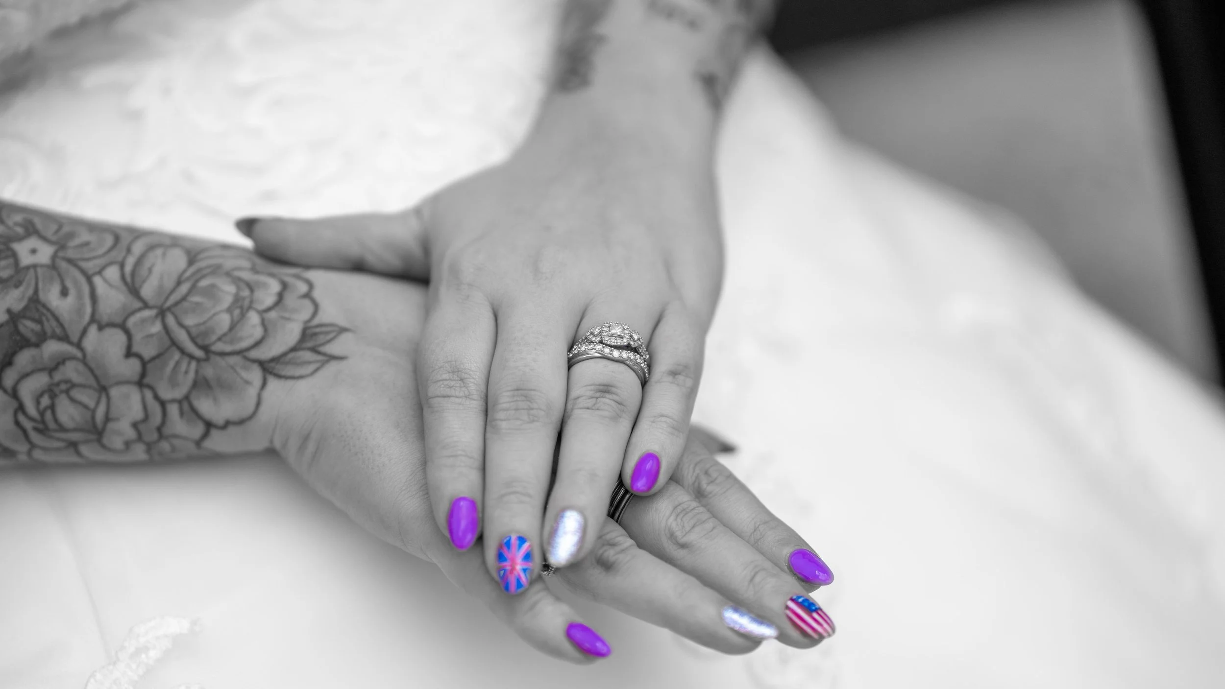 Close-up black and white photograph of a woman's hand with colorful nails and rings, resting on another person's hand with tattoos, on a white surface.