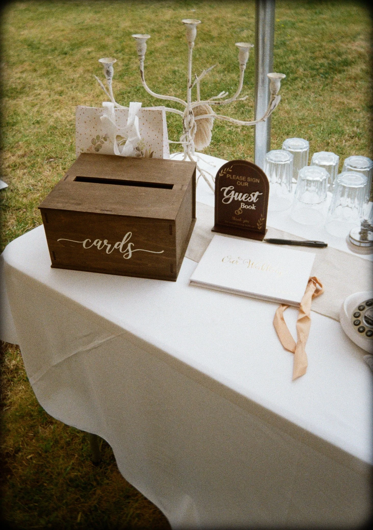 A white table with a guest book, a black pen, a decorative sign, a wooden box labeled 'cards', a gold and white chandelier, and upside-down glass cups on a white tablecloth, outdoors with grass in the background.