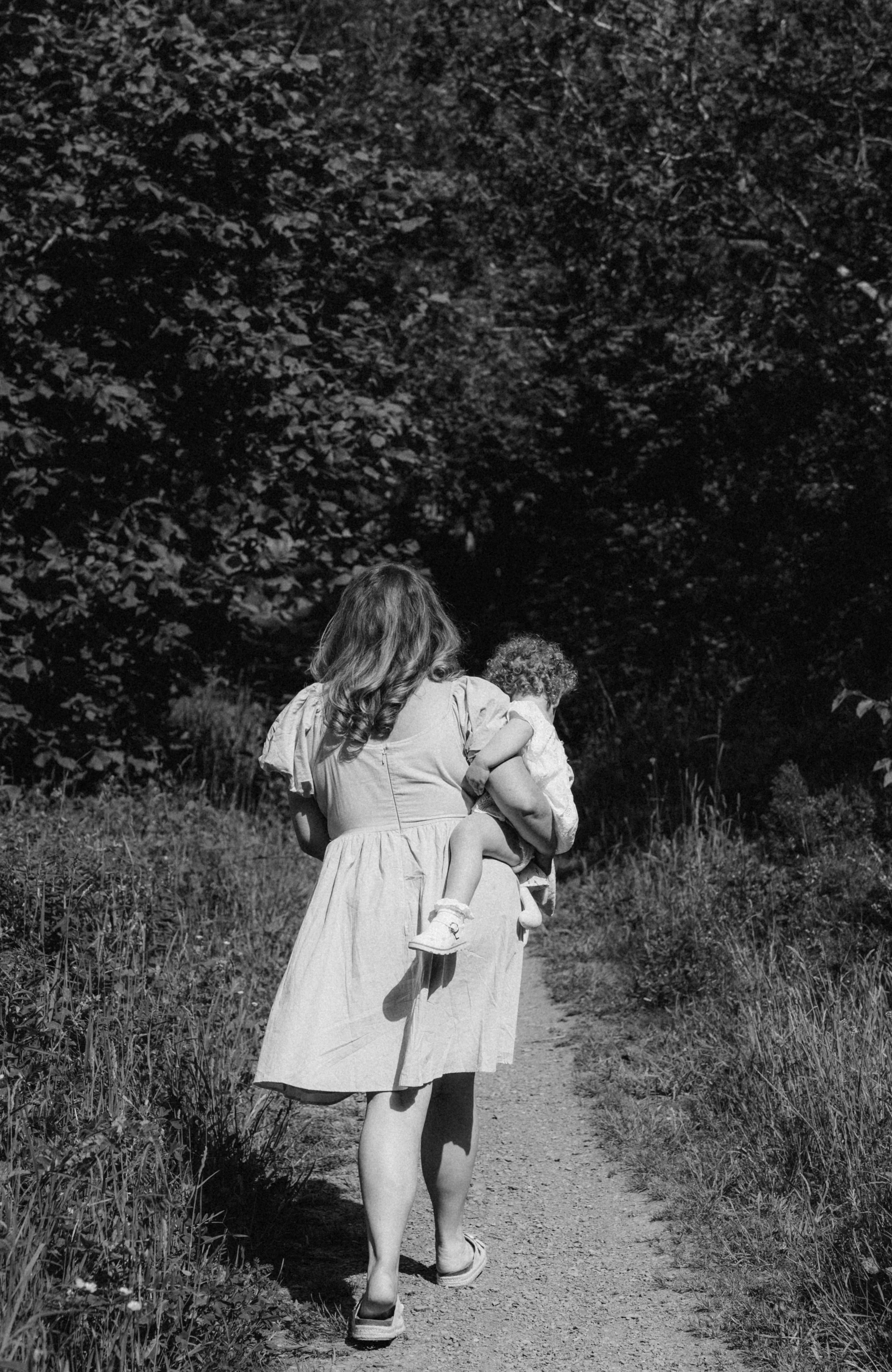 A woman in a dress walking on a dirt path while holding a young child in her arms, surrounded by tall grass and trees.