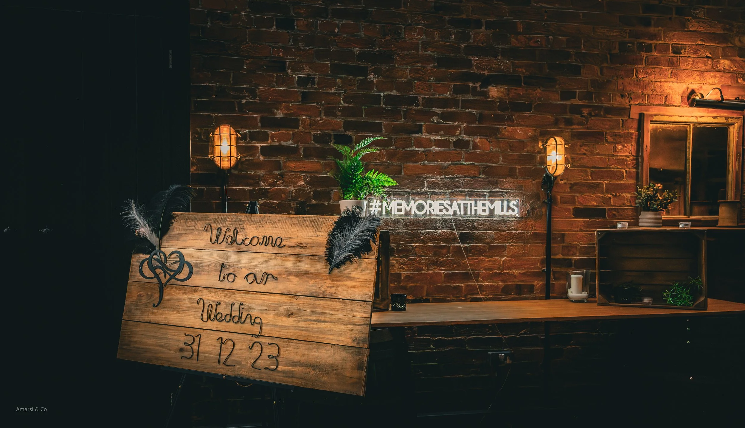 Wedding signboard with the message 'Welcome to our Wedding 31 12 23', decorated with feathers and a heart illustration, situated in front of a brick wall with potted plants, illuminated by warm lighting, with a neon sign '#MEMORIESATHEMLS' on the wal