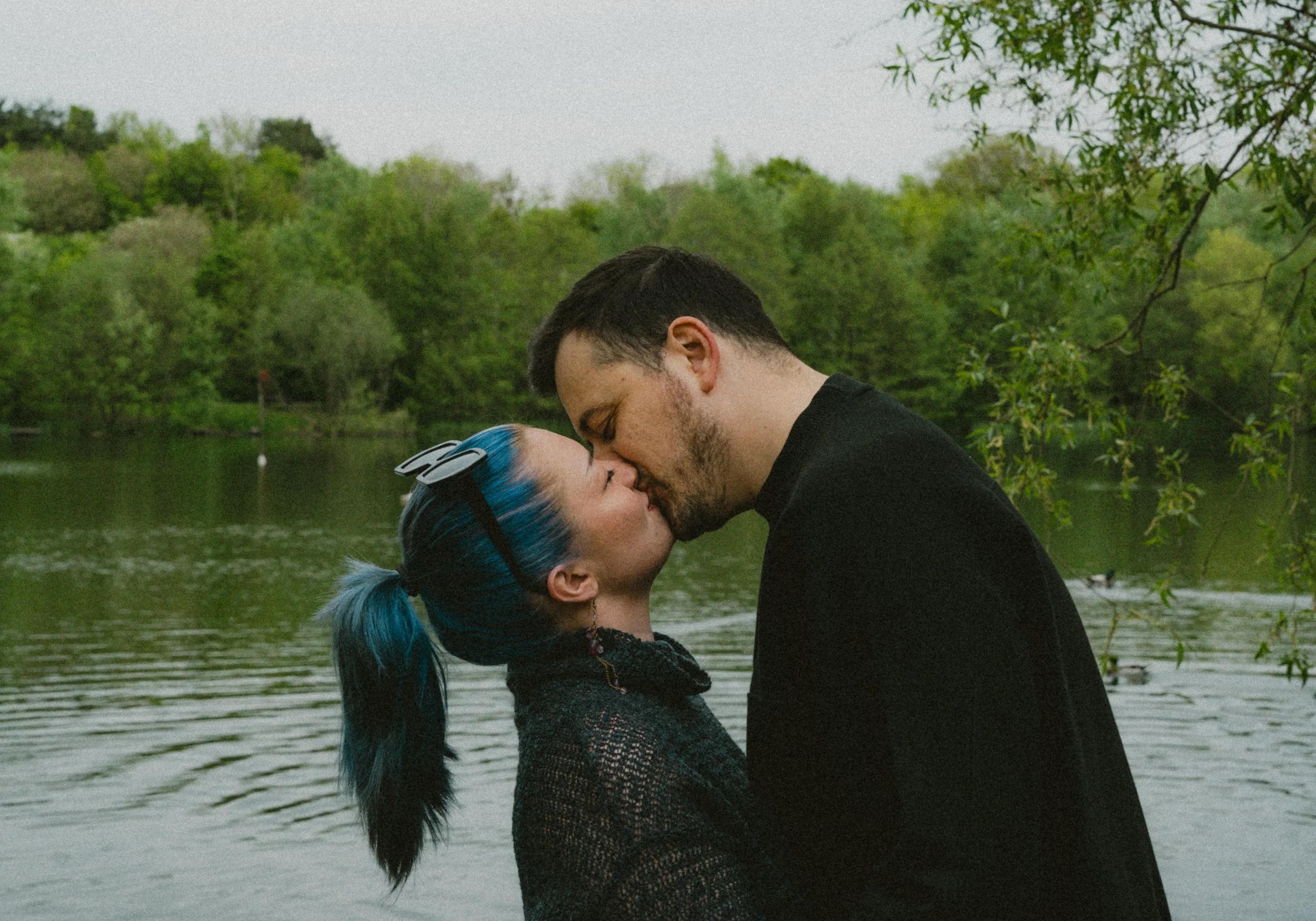 A couple kissing outdoors near a lake with green trees in the background.