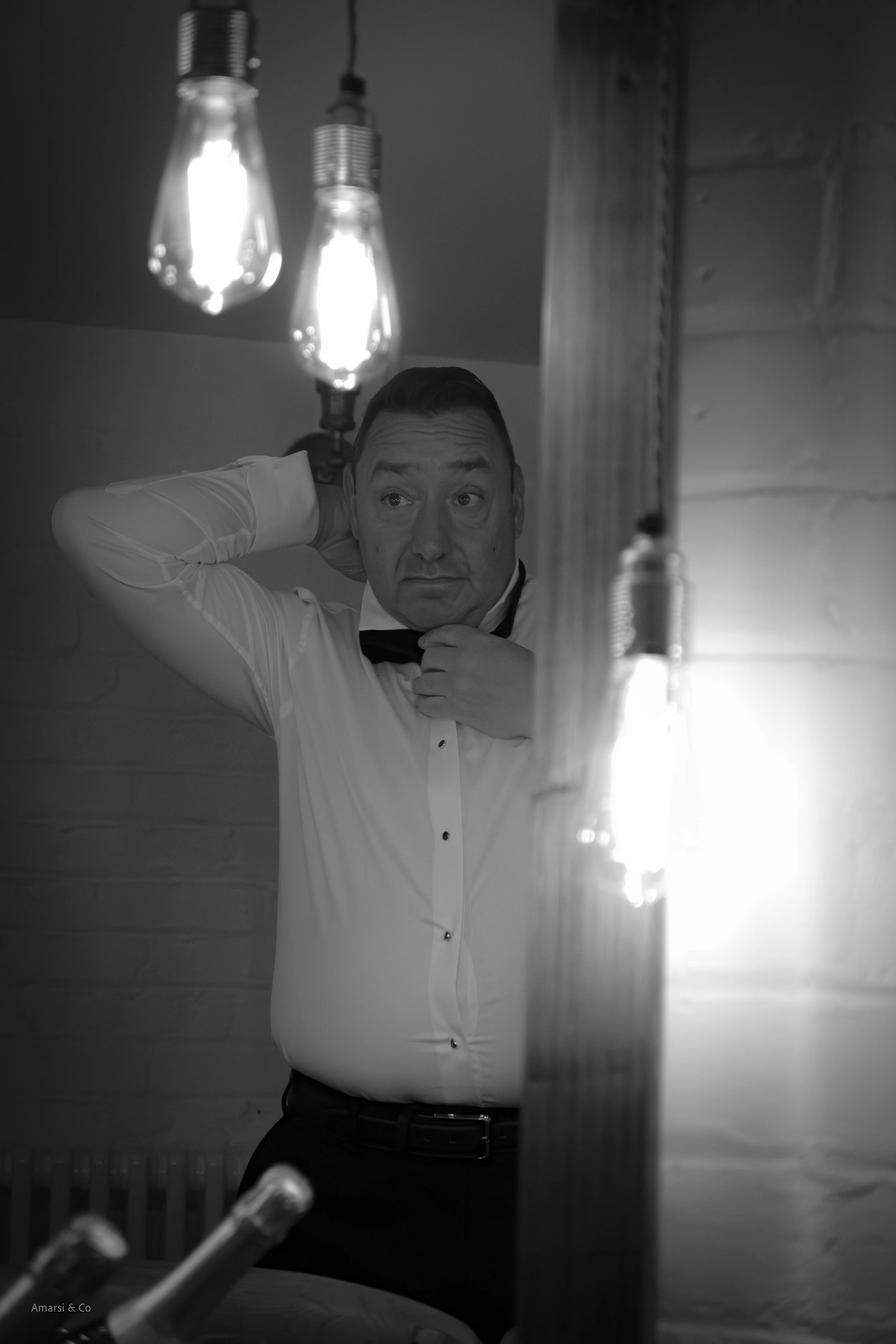 Man in a tuxedo adjusting his bow tie, looking into a mirror, with hanging light bulbs overhead.
