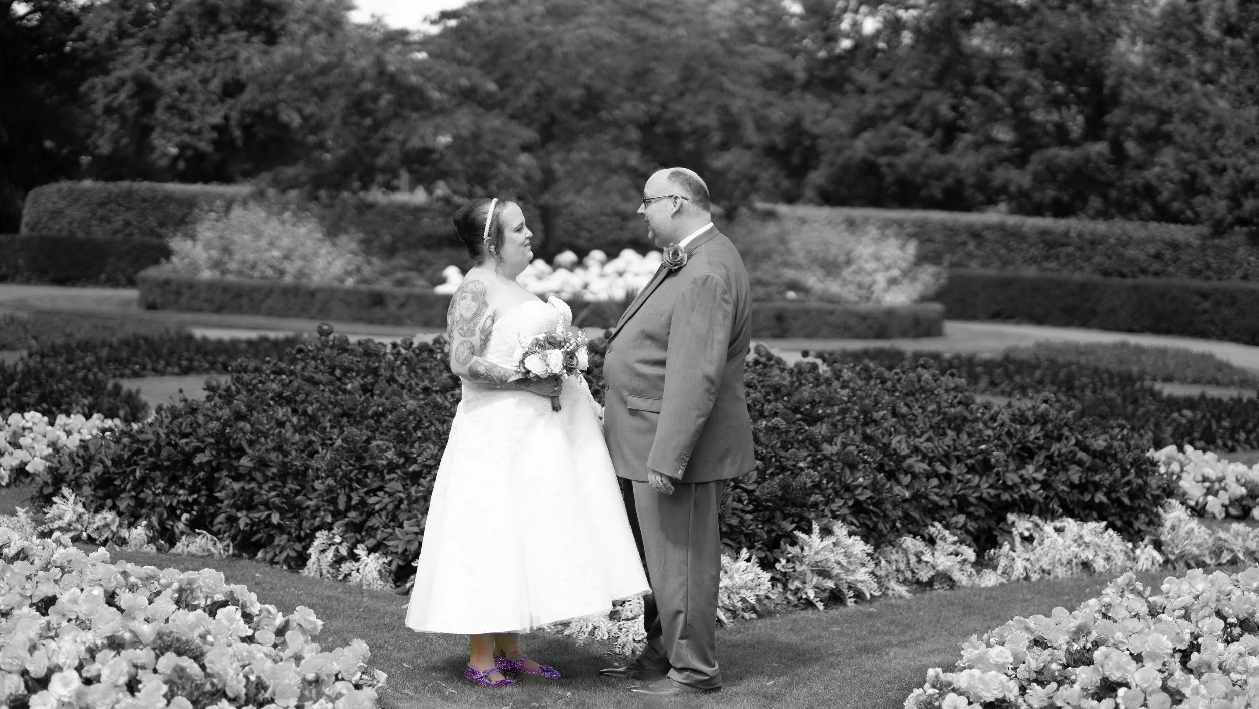 A bride and groom stand face to face in a garden, holding hands. The bride is wearing a white wedding dress and purple shoes, has tattoos on her arms, and is holding a bouquet. The groom is dressed in a suit with a boutonniere. The background feature