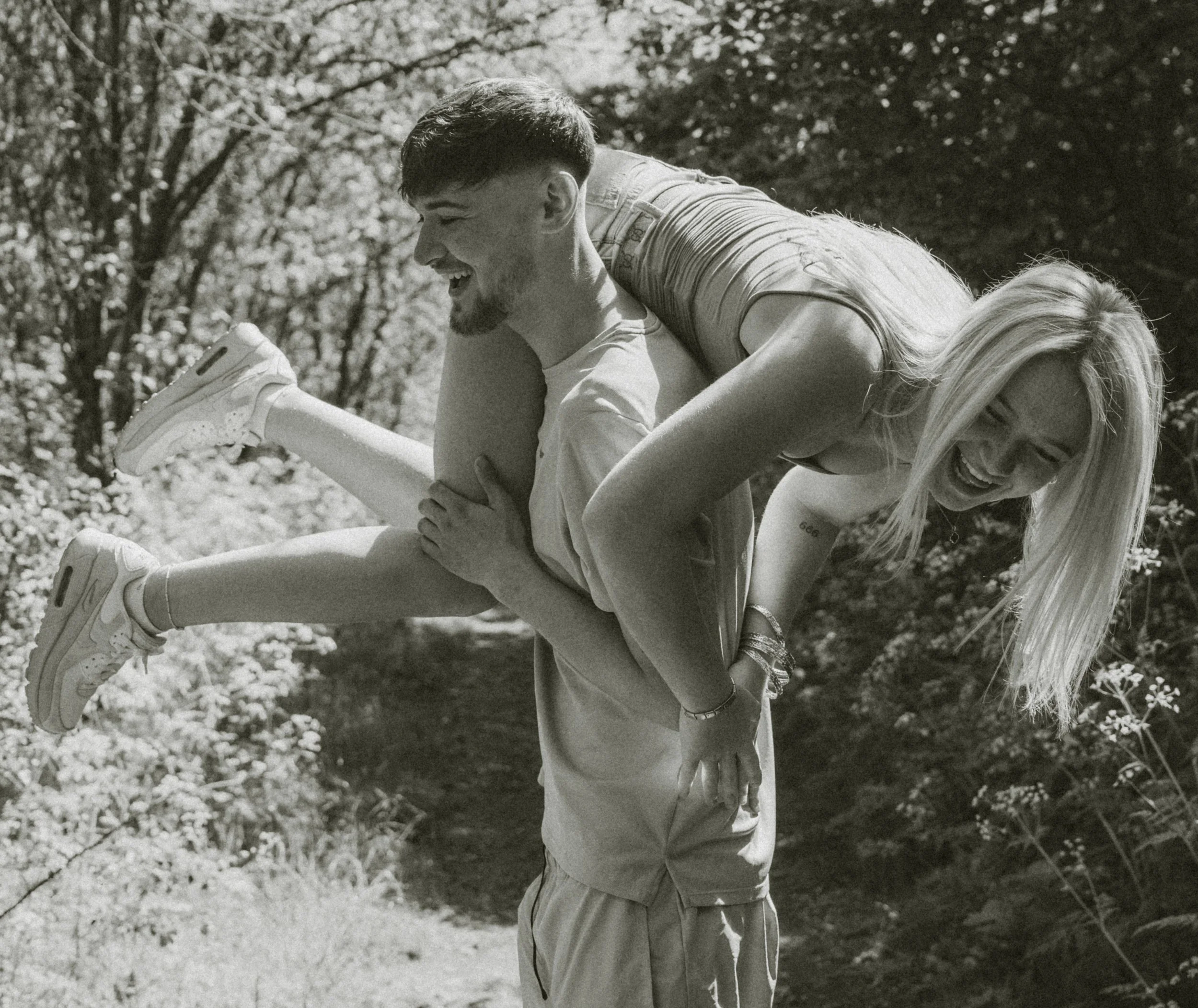 A man carrying a woman on his back outdoors. The man is smiling and looking down, while the woman is laughing with her head tilted back. They are surrounded by trees and natural scenery.