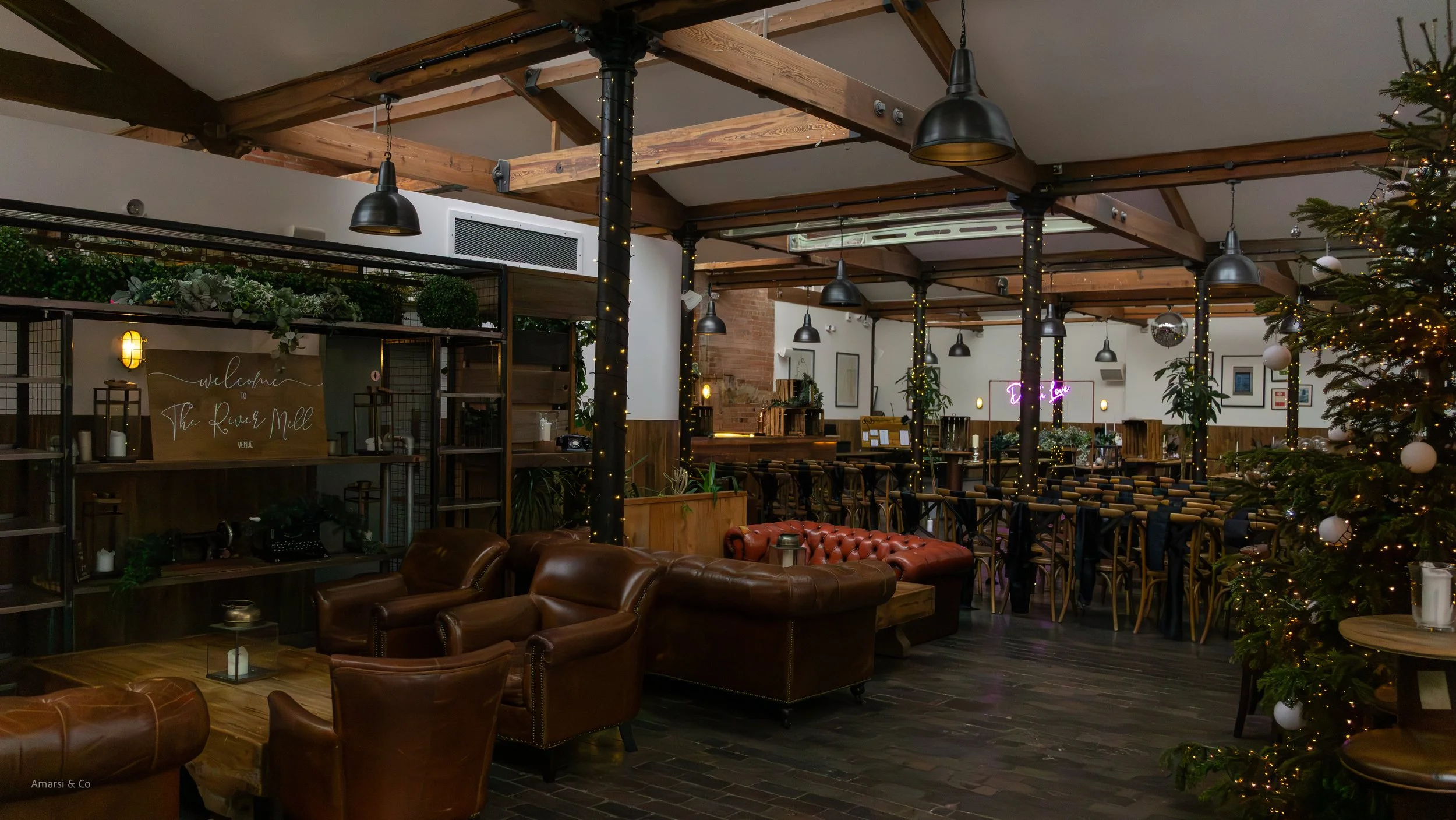 Interior of a cozy restaurant decorated for the holidays with a Christmas tree adorned with white ornaments and fairy lights, leather armchairs, a bar area with barstools, and warm lighting.