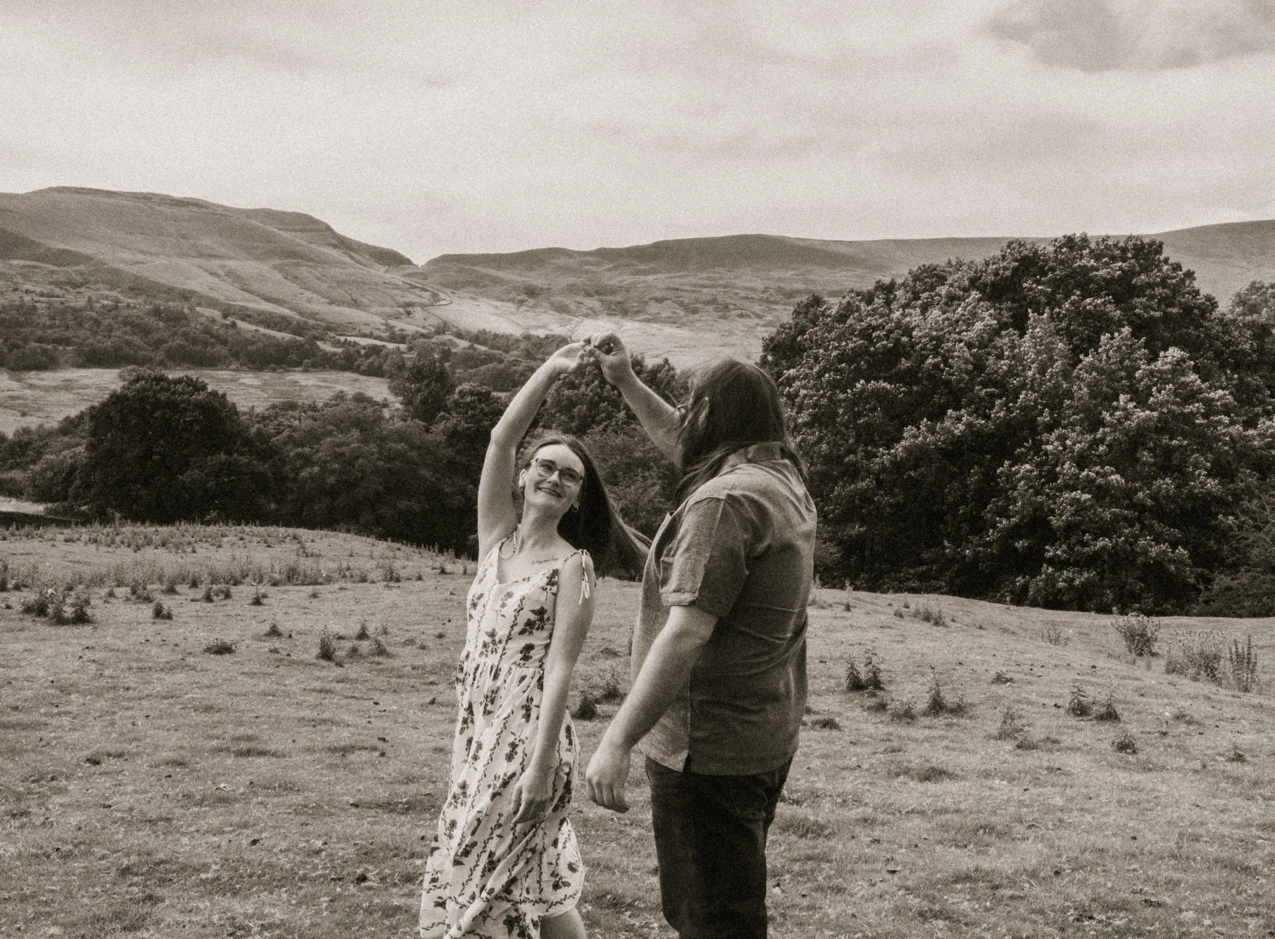 Two women dancing outdoors in a countryside landscape with hills, trees, and fields, in a black and white photo.
