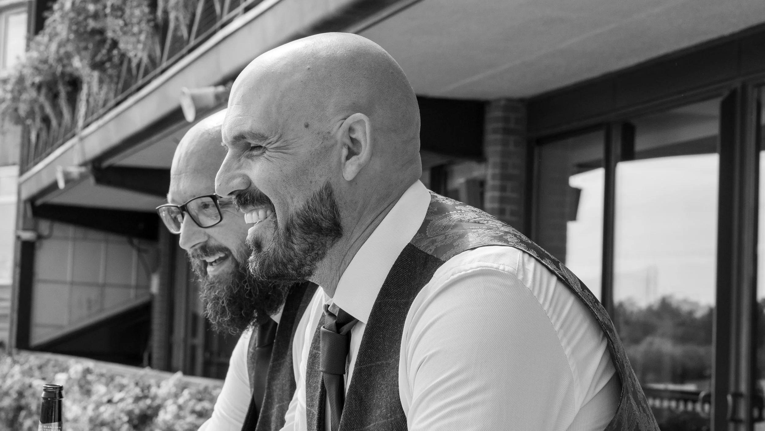Two men with shaved heads and beards, smiling and leaning close together outdoors, with a building and balcony in the background, in black and white, at Leicester Racecourse wedding.