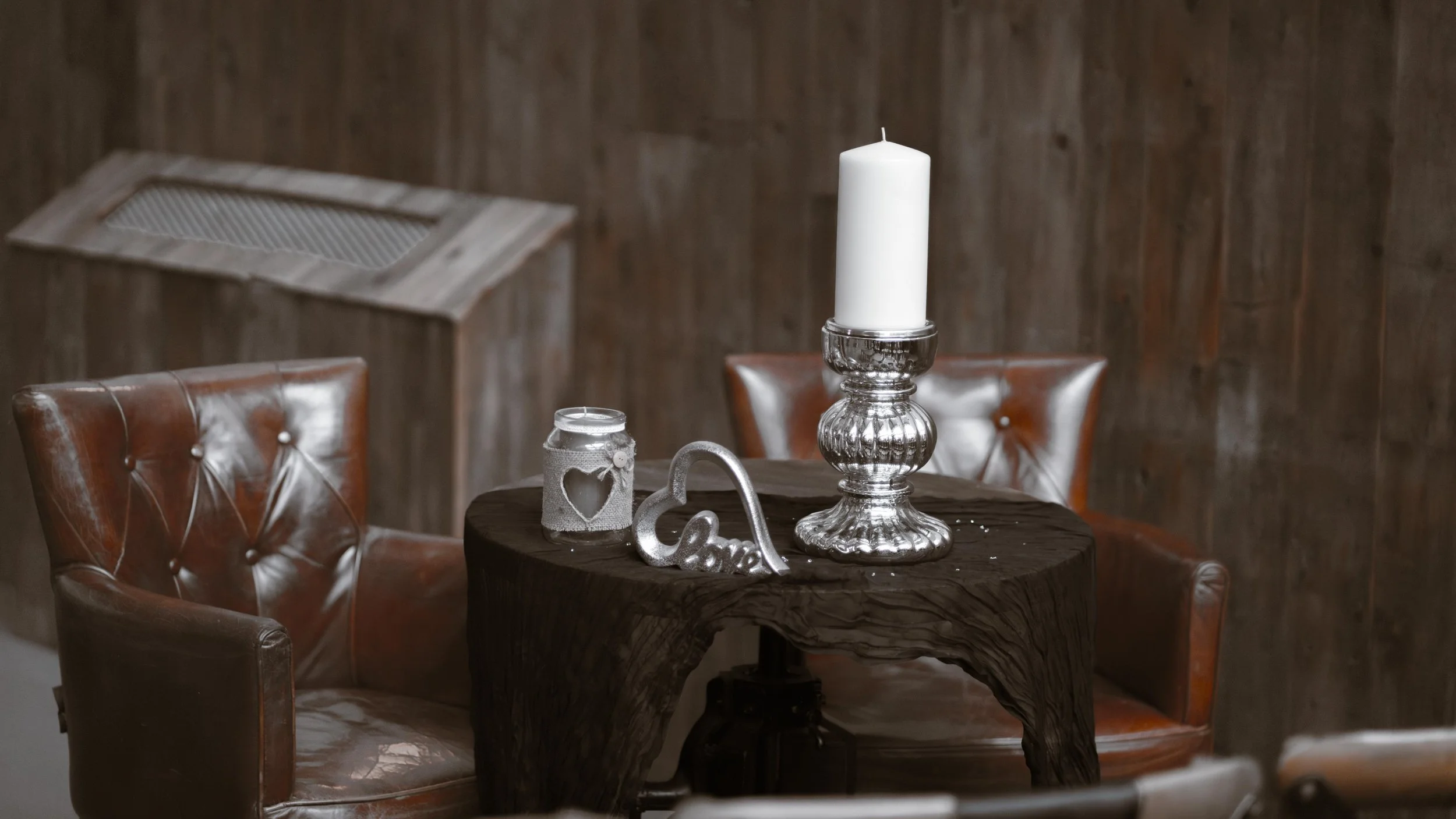 Cozy indoor setting with a rustic wooden wall, a round wooden table with a silver candlestick holding a white candle, decorative elements including a small jar with ribbon and a heart-shaped ornament, and two leather armchairs.
