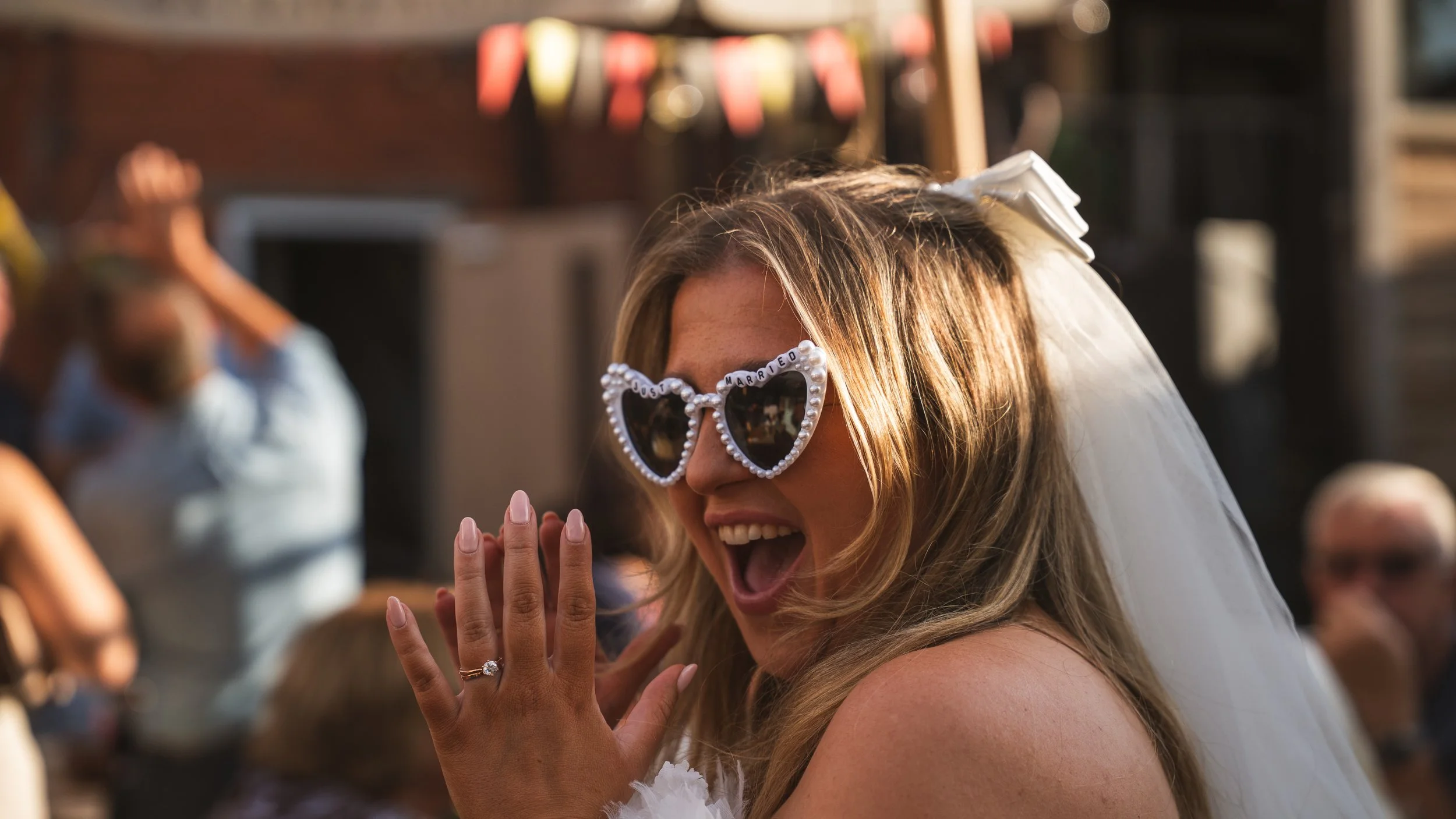 A woman with blonde hair wearing heart-shaped sunglasses with the words "Just Married" on them, smiling and holding her hands together, possibly during a celebration or wedding in Chesterfield.