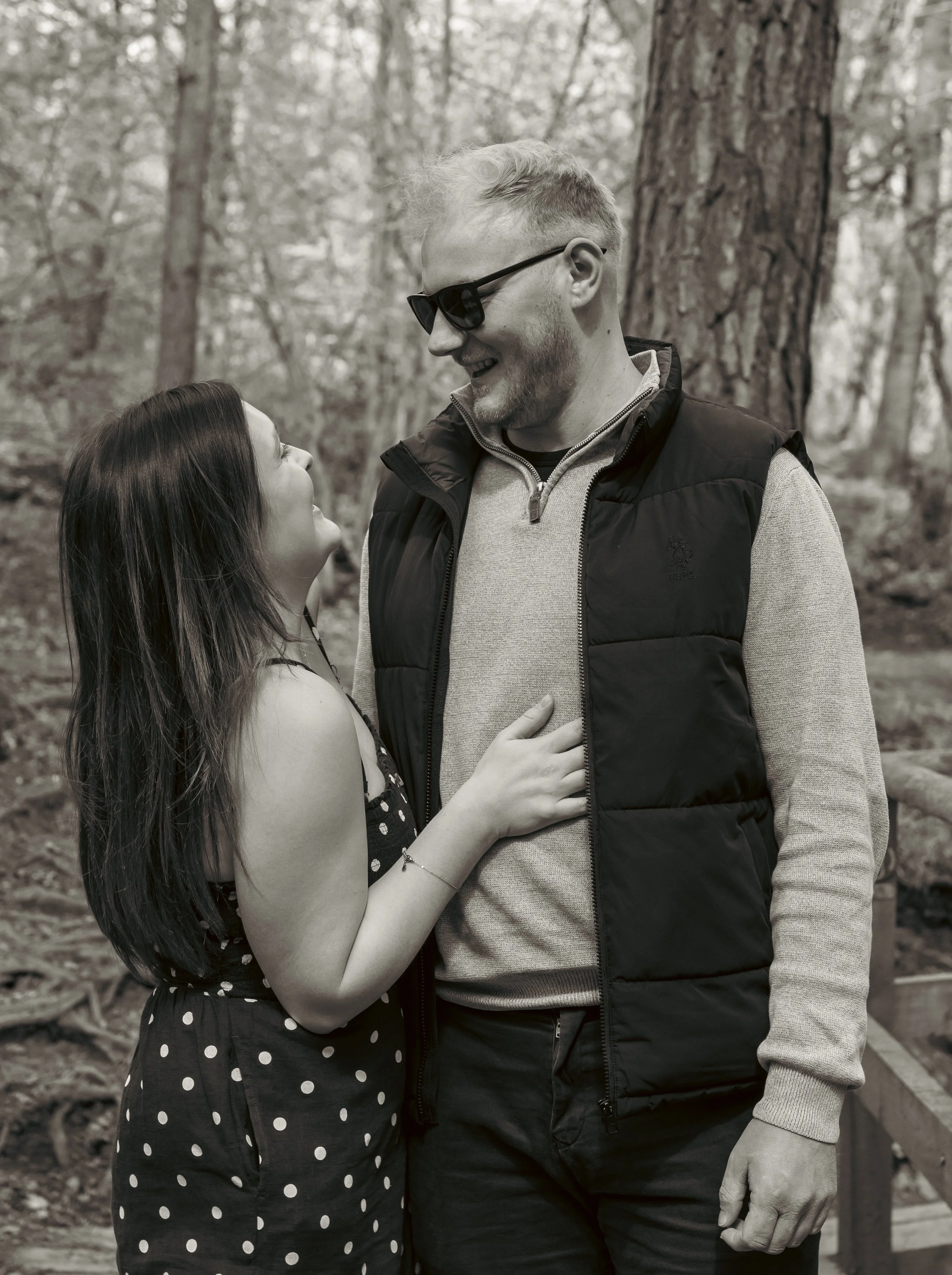 A young woman and an older man are standing close together outdoors in a forest, smiling and looking at each other, with the woman touching the man's chest.