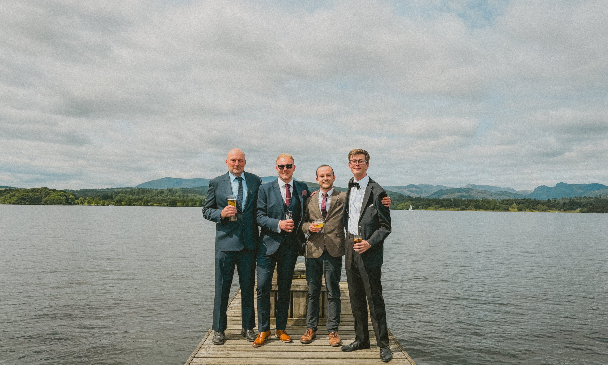 Four men in suits standing on a wooden dock by a lake, holding drinks, with mountains and a cloudy sky in the background. Low wood bay, the Lake District.