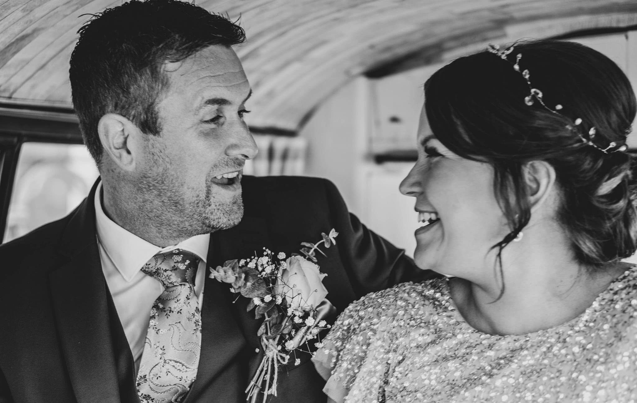 Black and white photo of a smiling man and woman looking at each other, dressed in formal attire, inside a vehicle.