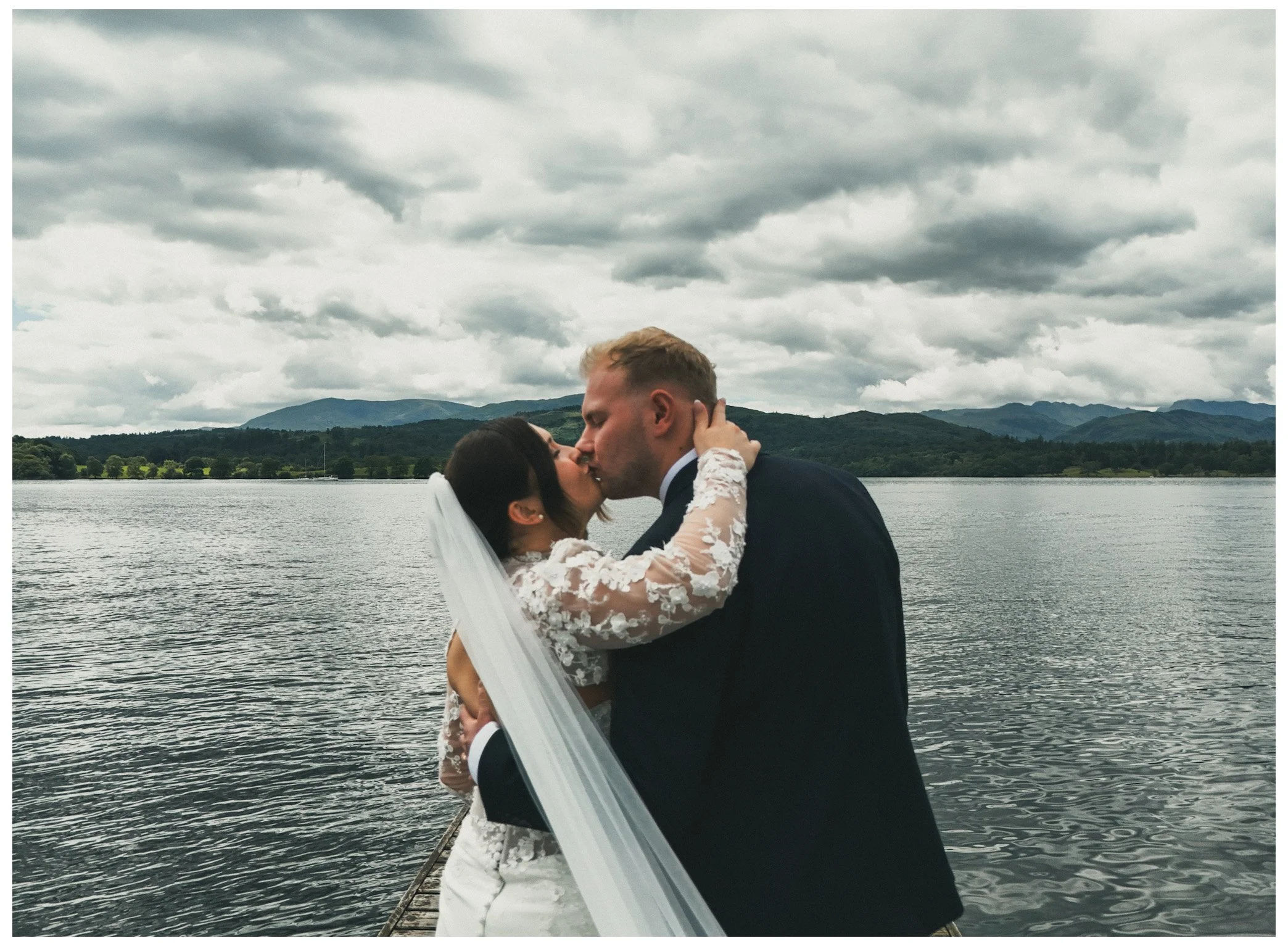 A bride and groom kissing outdoors on a dock with a lake and mountains in the background, under cloudy skies.