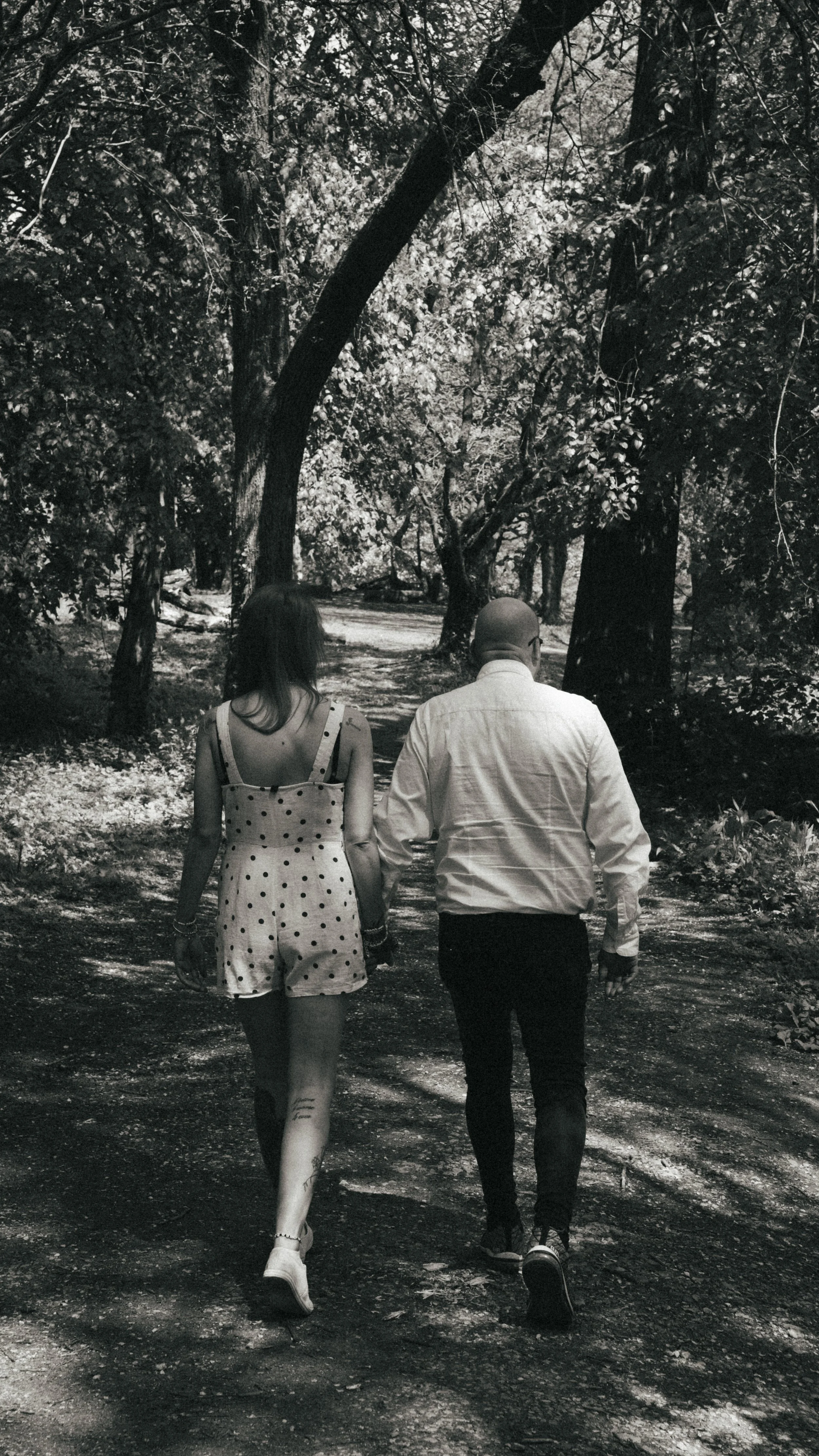 A black and white photo of a man and woman walking hand in hand on a wooded path.