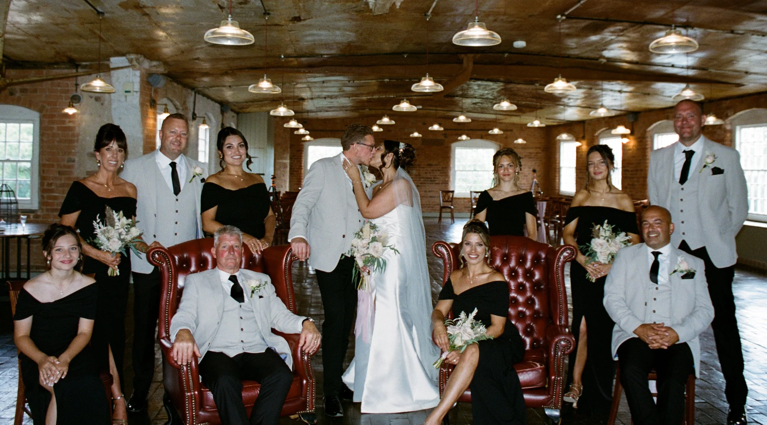 A group of people is gathered for a wedding, including the bride and groom in the center, surrounded by friends and family, in an indoor rustic venue with wooden ceiling panels and brick walls.