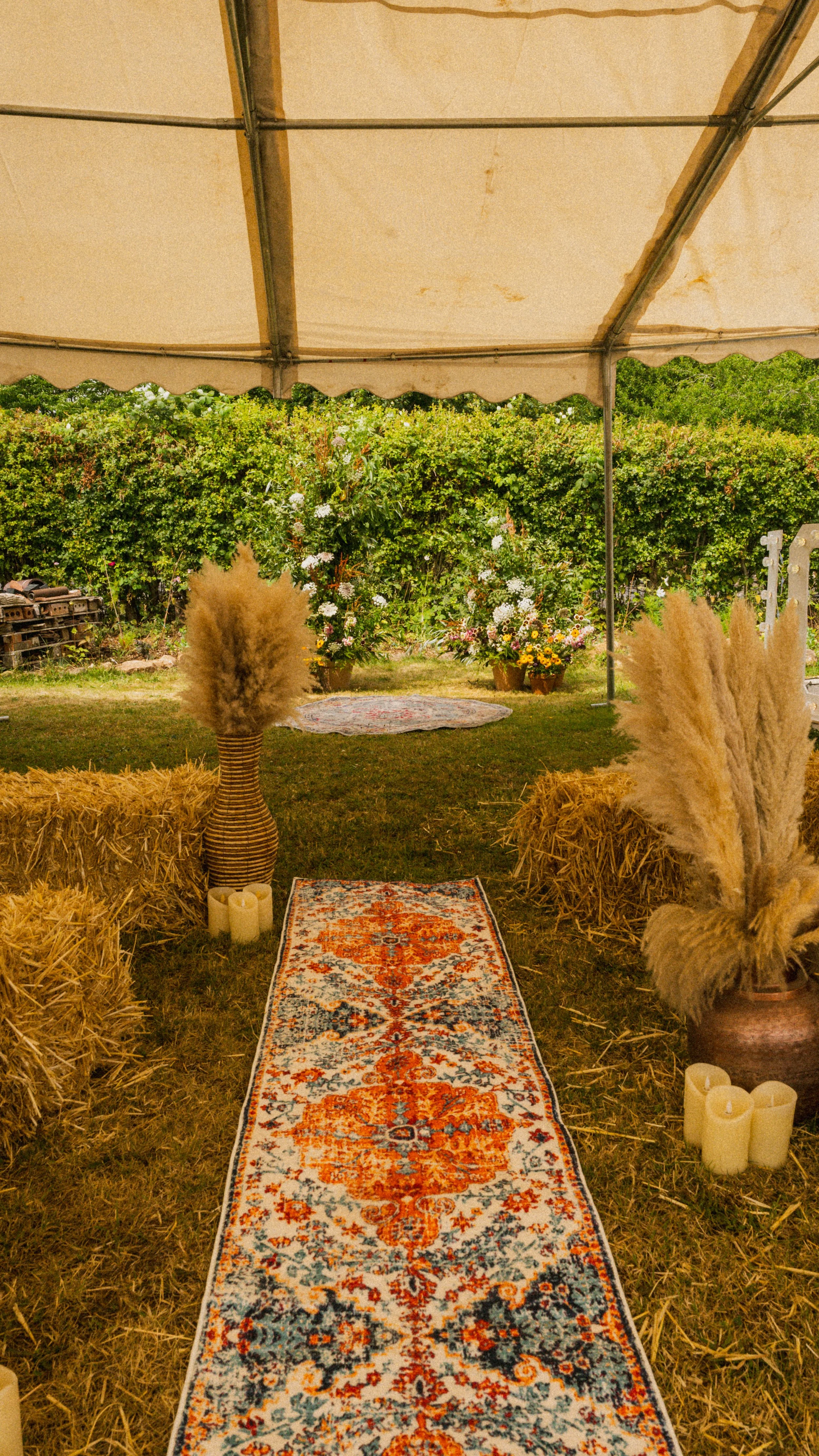 A decorated outdoor tent with hay bales, pampas grass in vases, candles, and a colorful rug on the grass, with greenery and flowers in the background.