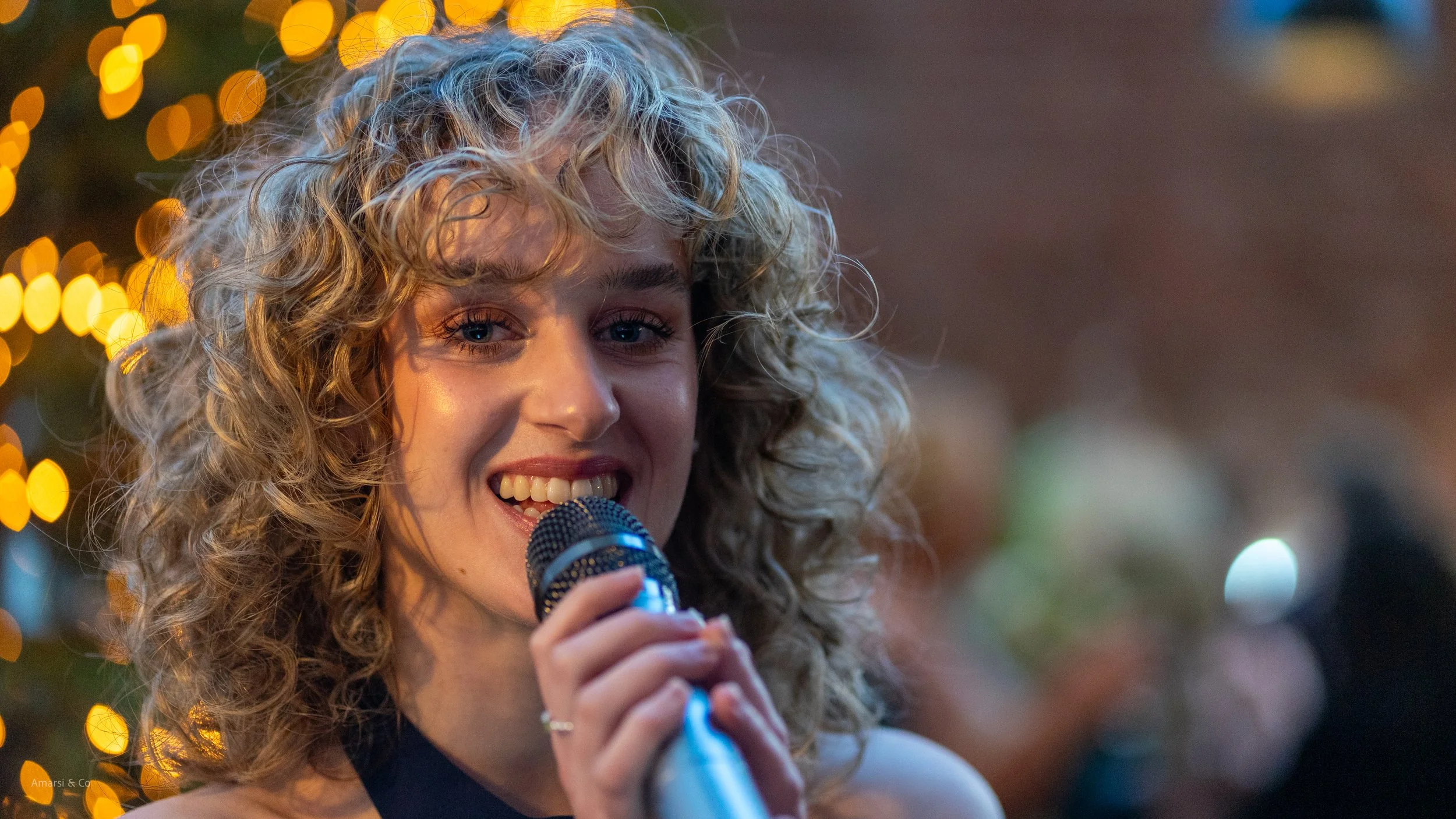A woman with curly blonde hair smiling and holding a microphone at an event with warm, festive lighting in the background.
