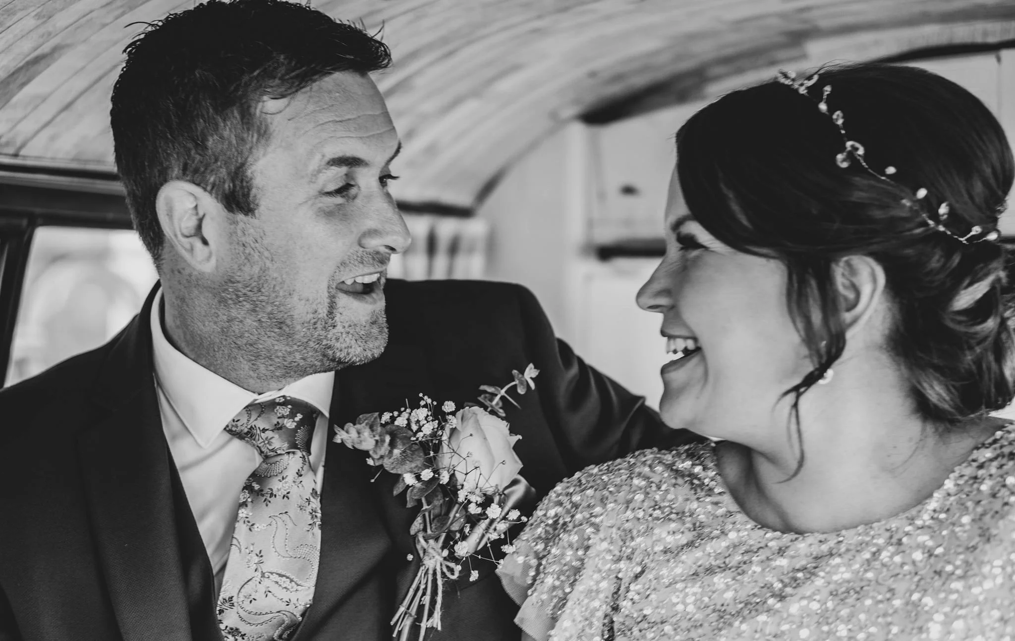 Black and white photo of a man and woman in wedding attire sharing a joyful moment, smiling at each other, with the man wearing a suit with a boutonniere and the woman in a sequined dress with a decorative hairstyle.
