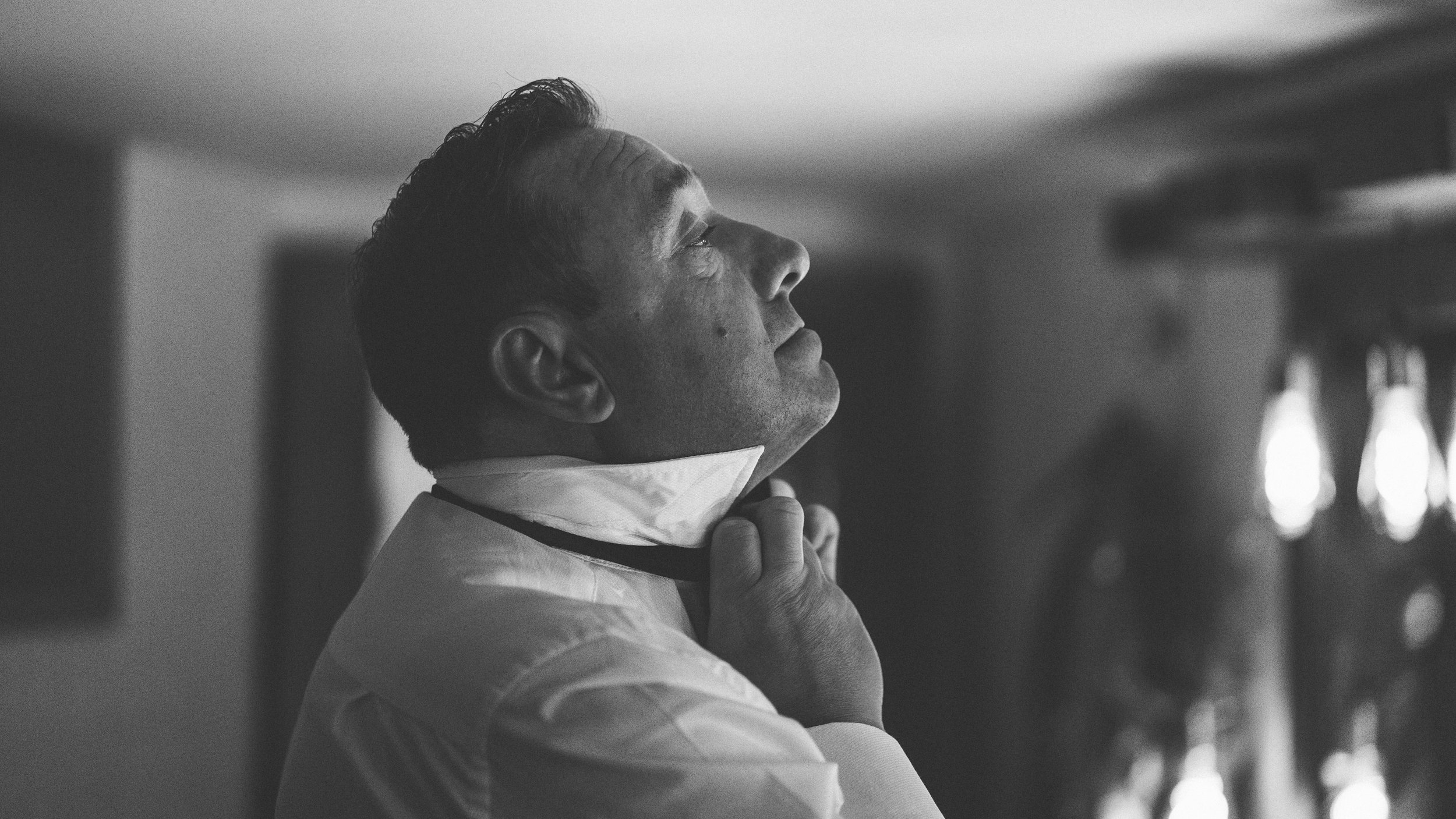 A man adjusts his tie while looking up, with light illuminating his face and focusing in the box of his face.