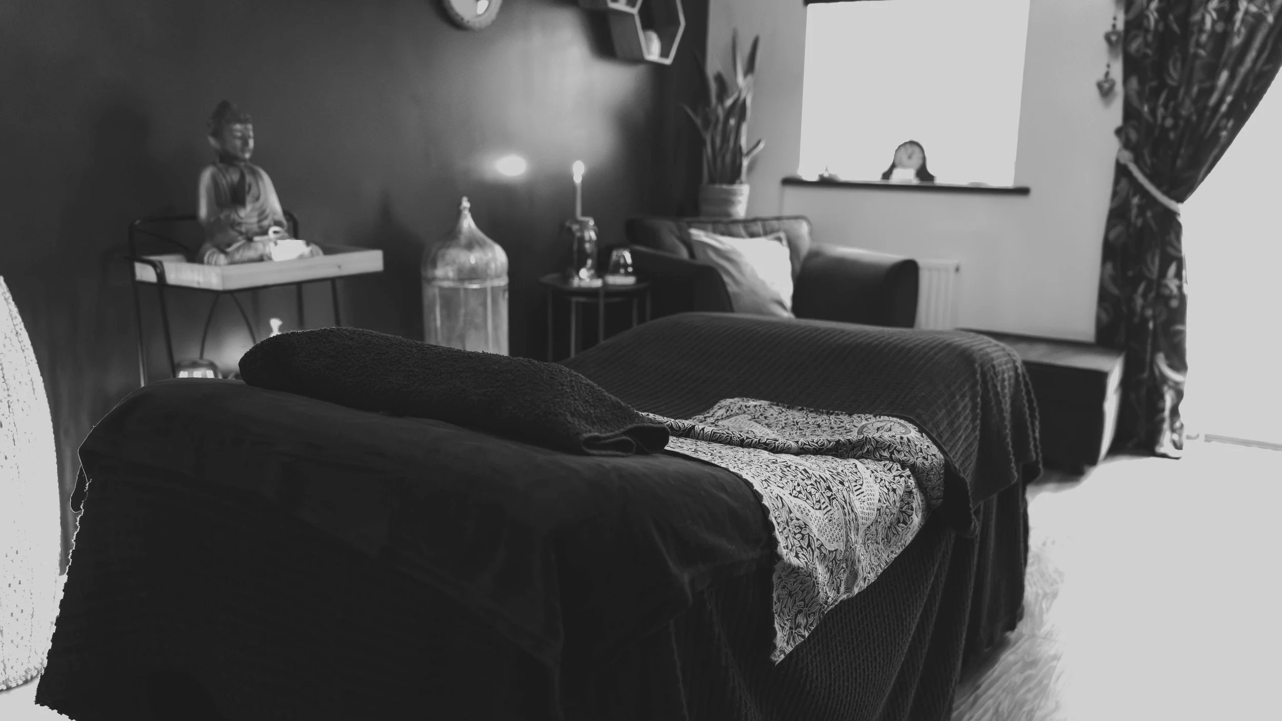 A cozy living room with a sofa covered with a dark blanket in the foreground, a side table with decorative items including a large glass bottle and a lit candle, a cushioned armchair, potted plants near the window, and a curtain with a floral pattern