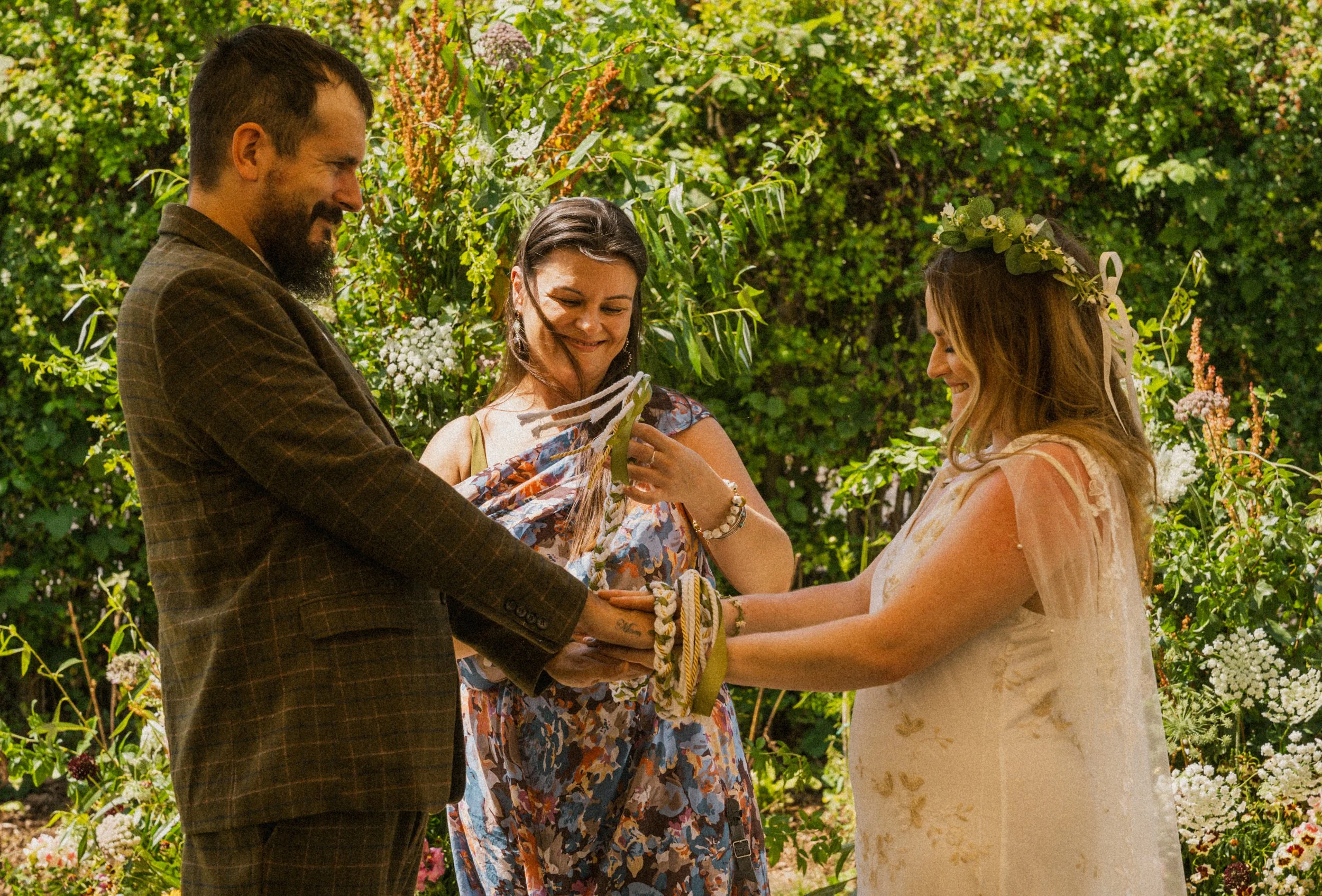 A couple gets married outdoors with a woman holding their hands during the ceremony, surrounded by lush green bushes and colorful flowers. At Rhubarb farm, Langwith