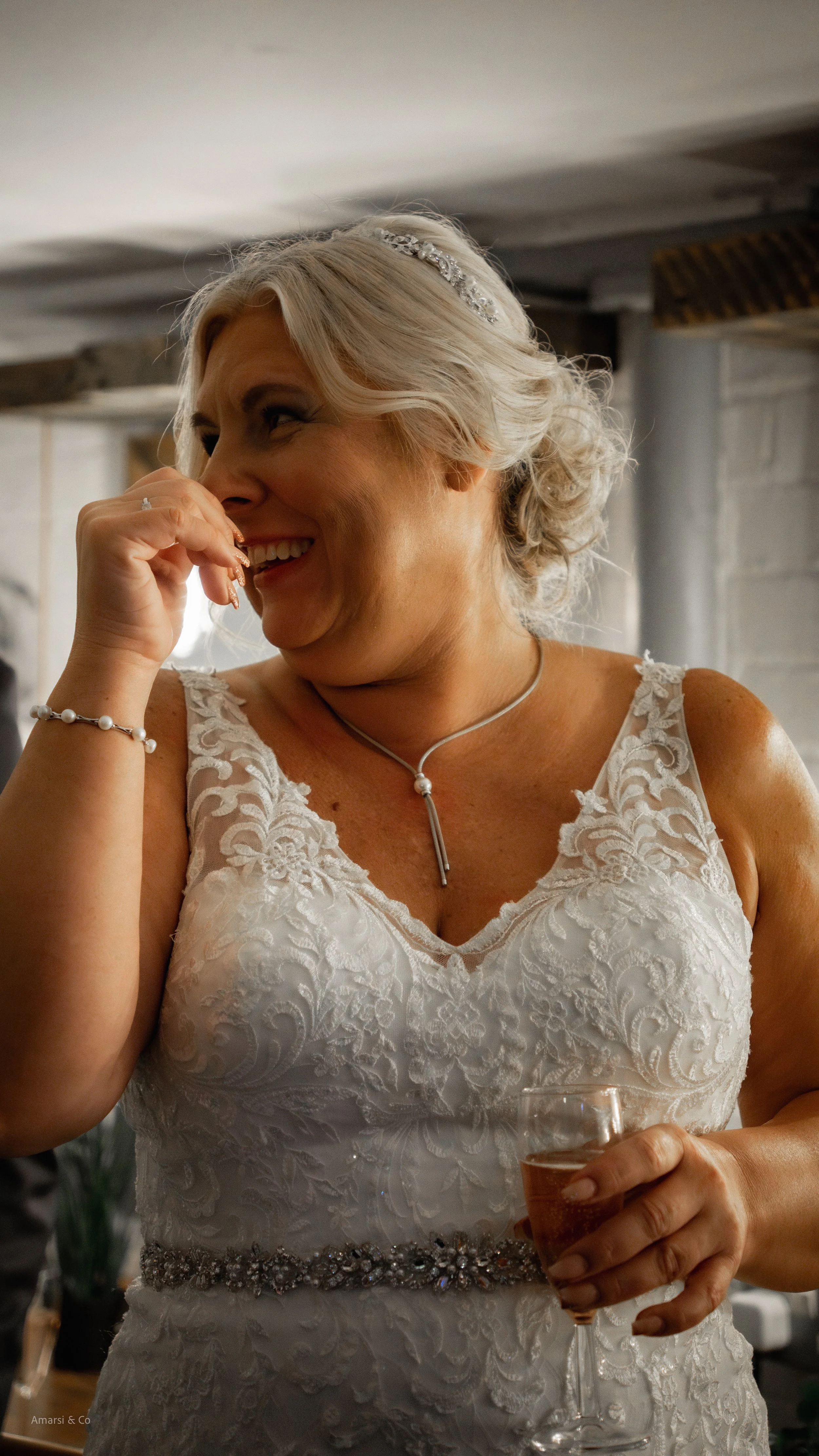 A woman in a wedding dress smiling and laughing, holding a glass of champagne, with her hand near her face and wearing jewelry. at the west mill derby