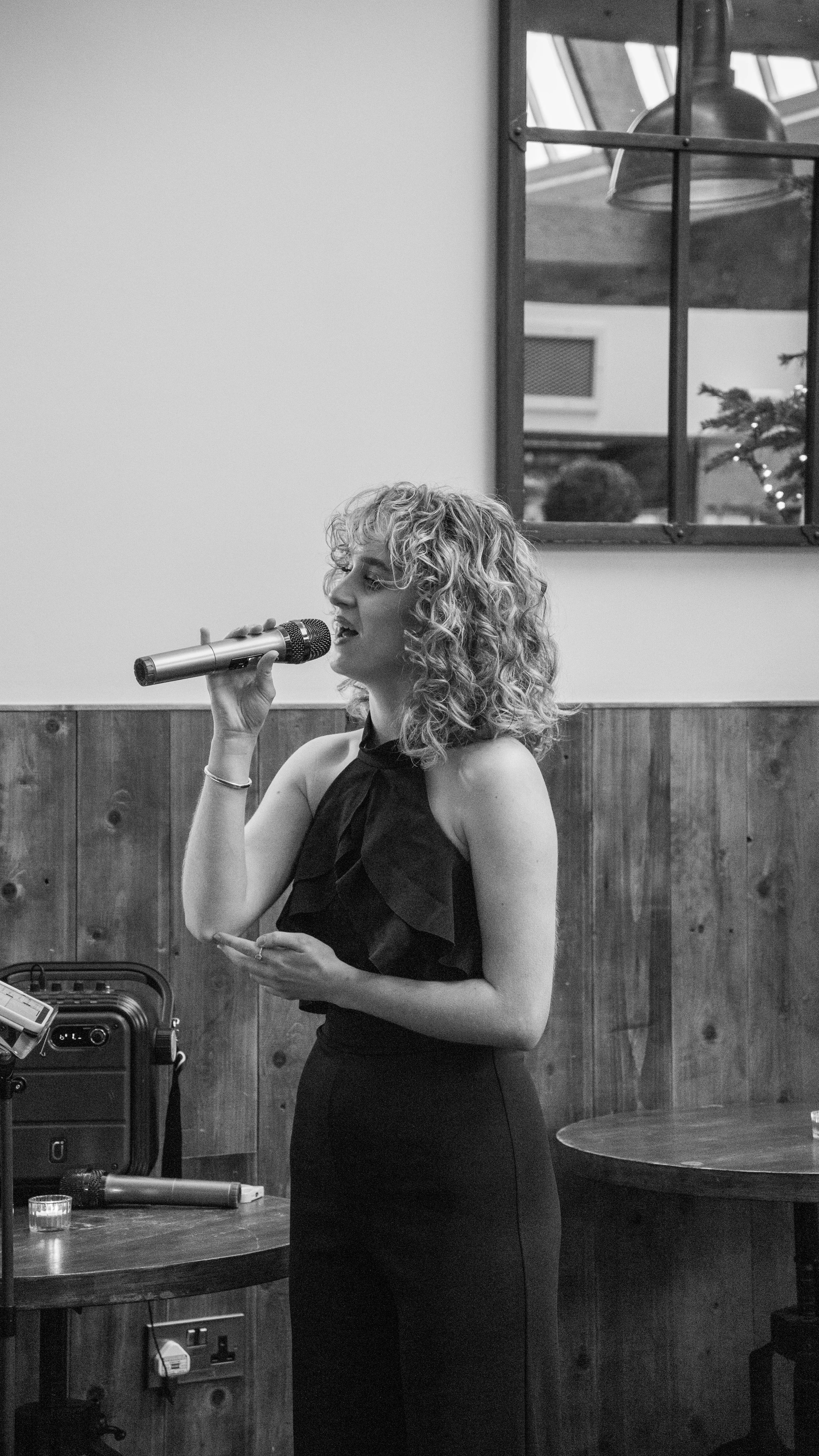 A woman with curly hair singing into a microphone in an indoor setting, with a window in the background and various items on a table nearby.