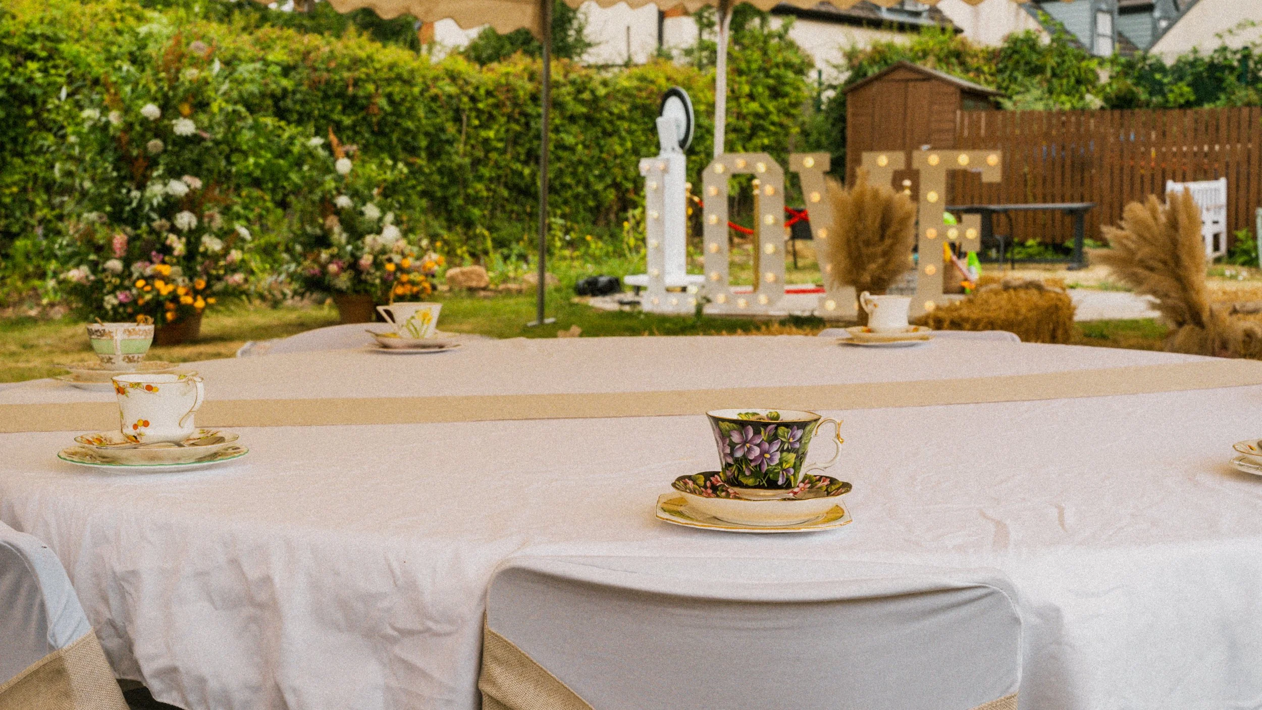 Decorative tea cups and saucers set on a white tablecloth in an outdoor garden, with a background of blooming flowers, greenery, and outdoor furniture.