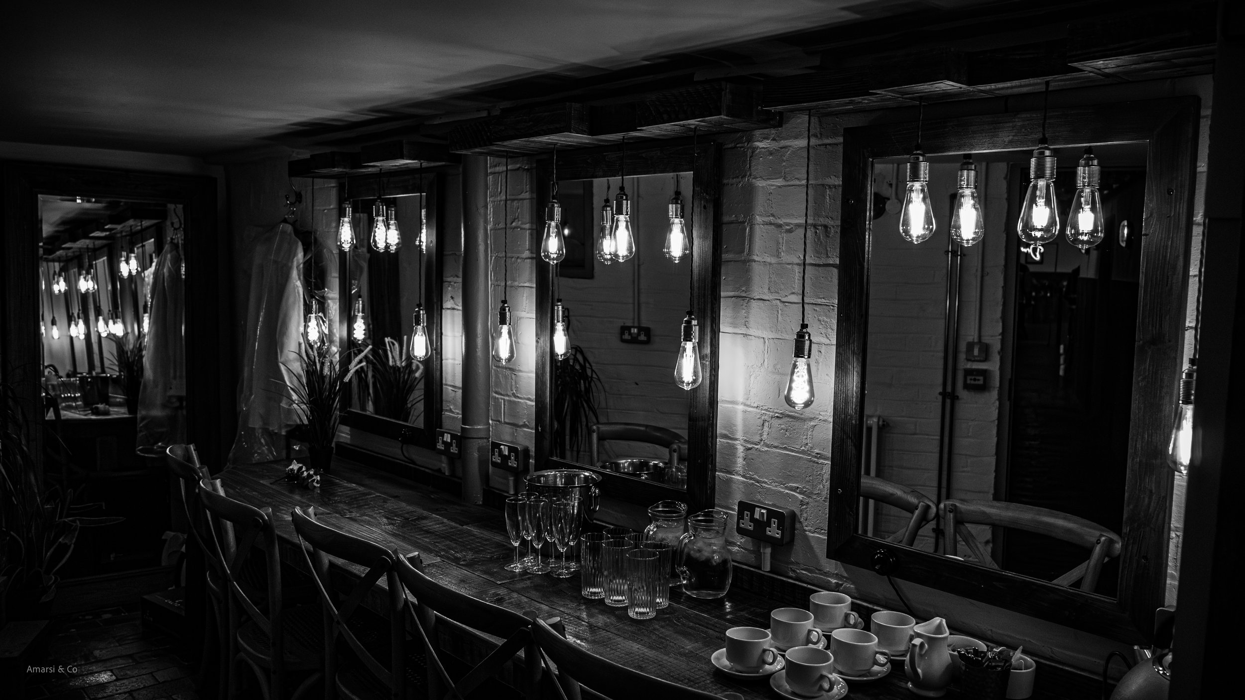 Interior of a kitchen or dining area with hanging Edison-style bulbs, a wooden table set with glasses and cups, and mirrors reflecting the room.