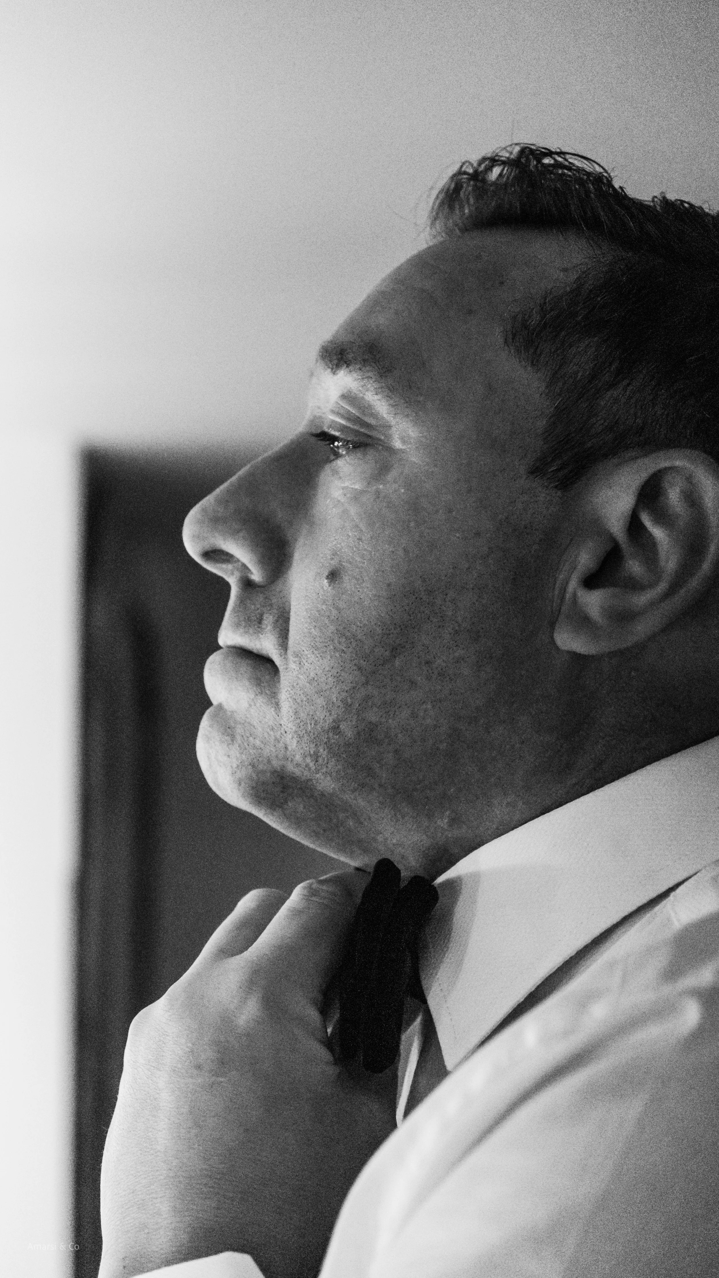 Close-up of a man adjusting a bow tie in front of a mirror, wearing a tuxedo, captured in black and white. at the west mill, derby