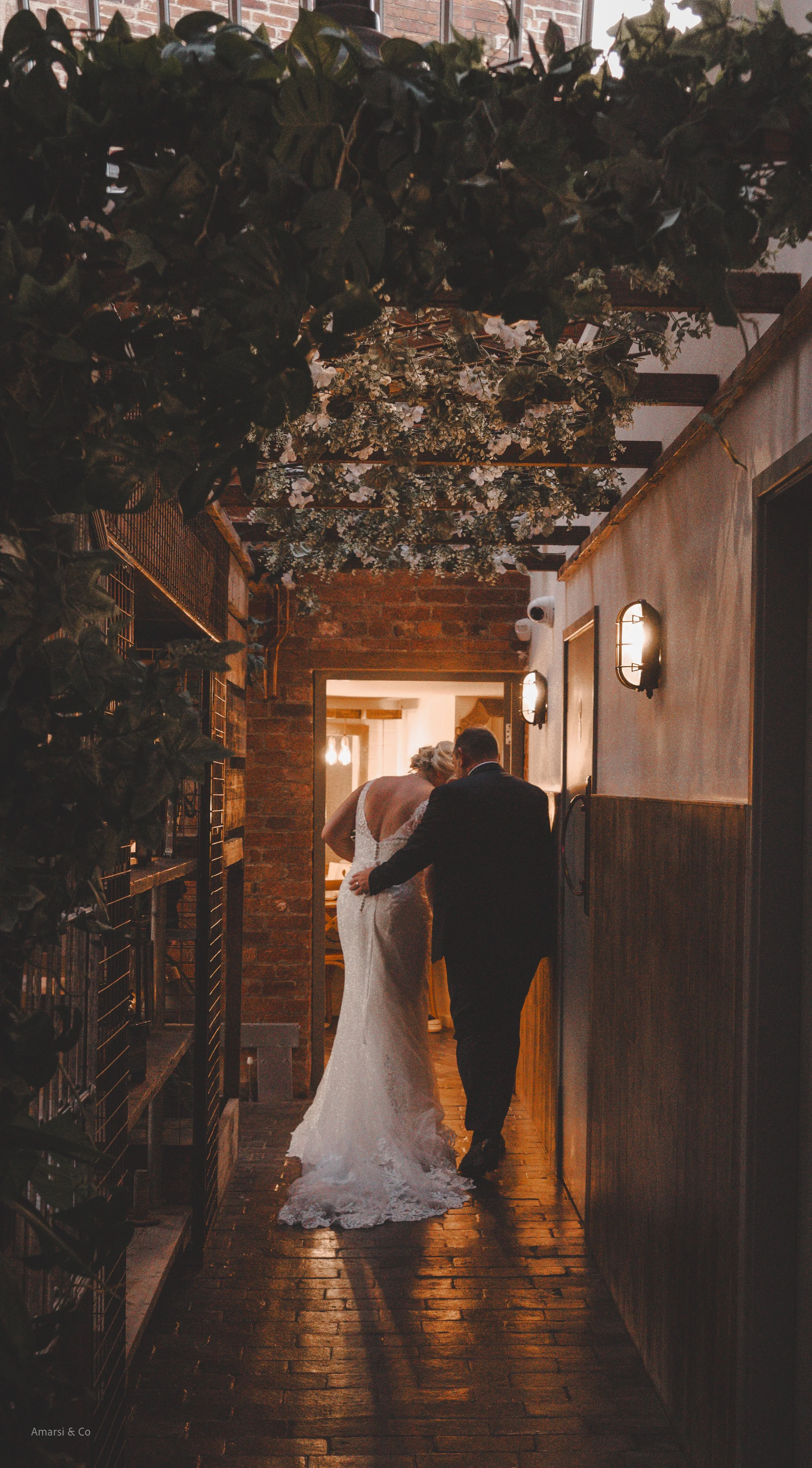A bride and groom walk hand-in-hand down a rustic hallway decorated with floral arrangements and warm lighting, with exposed brick walls. the river mill derby