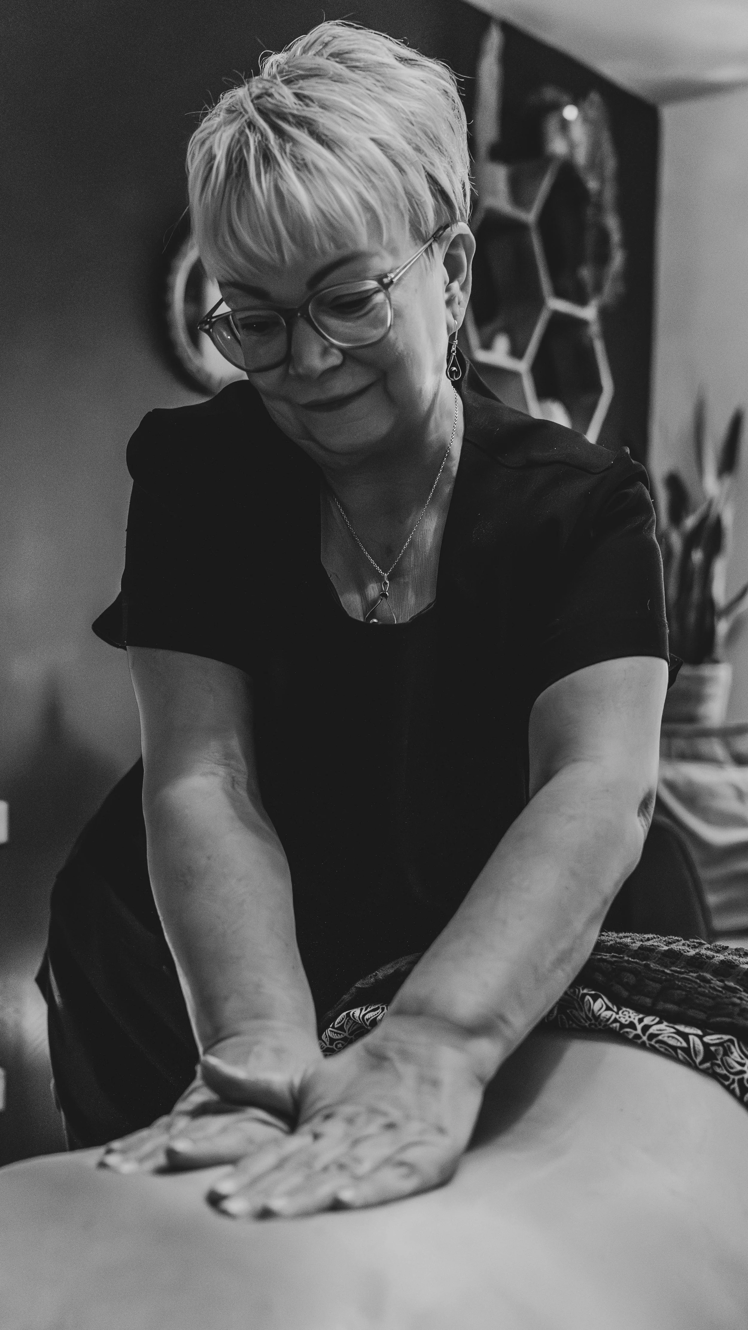 A woman with short, light-colored hair, glasses, and earrings leans over and places her hands on a person's body, possibly during a massage or health-related activity, with a hexagonal wall decor in the background.