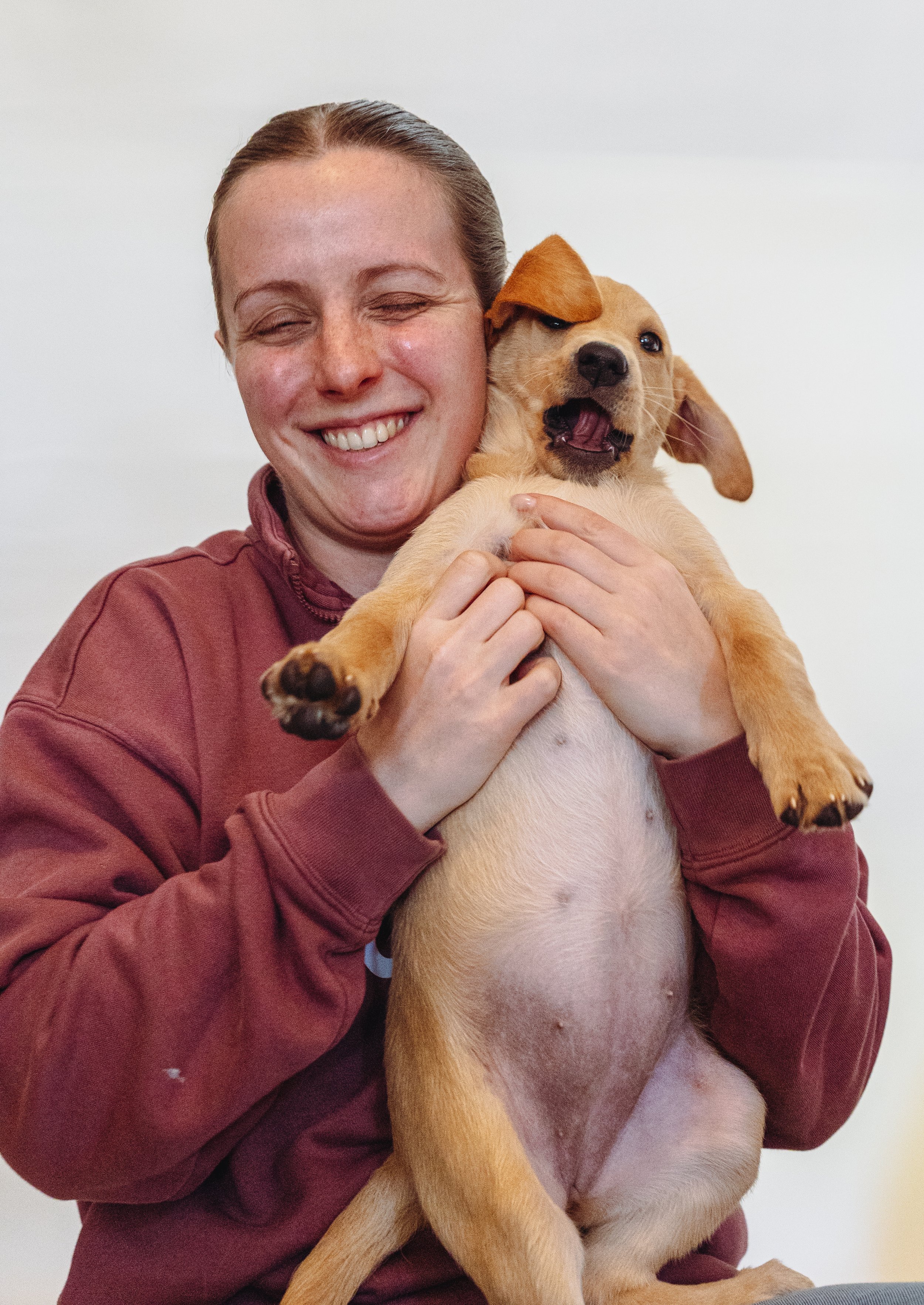 A woman with a big smile holding a light brown puppy with floppy ears and a happy expression, against a plain white background.