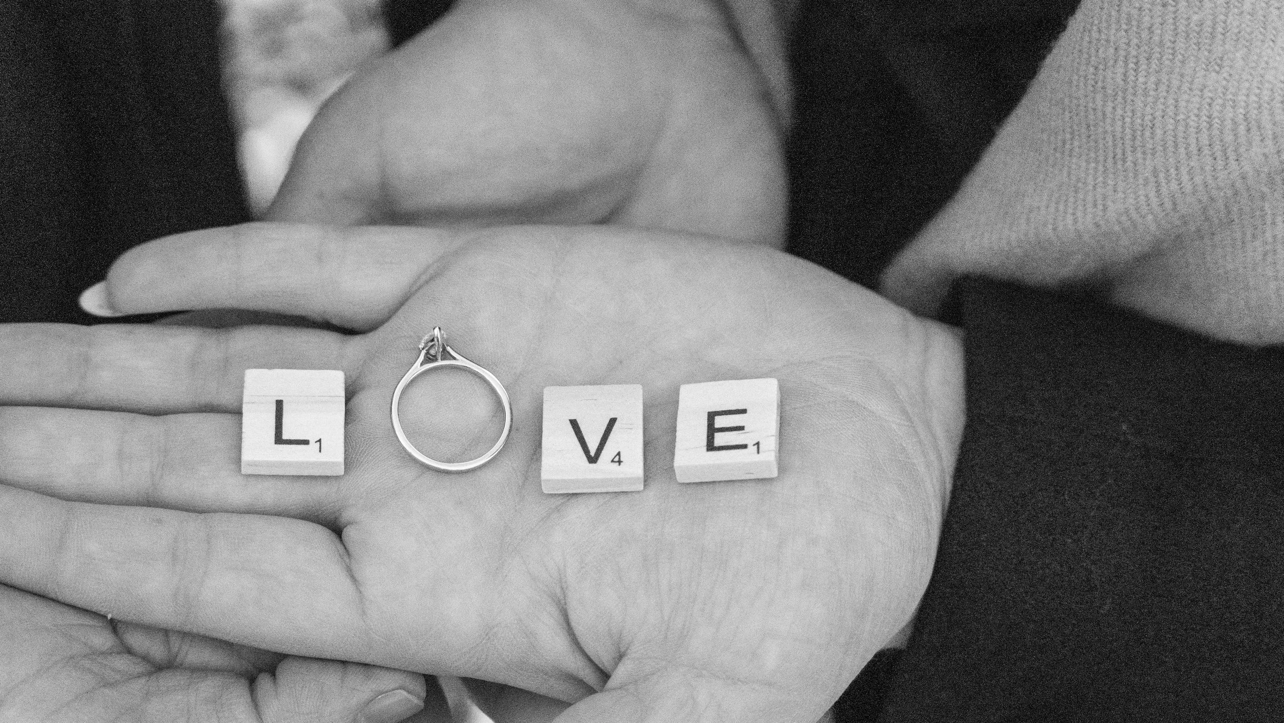 A close-up of two hands holding a small ring, with Scrabble tiles spelling out "LOVE" and a wedding band on the ring finger. The background is slightly blurred.
