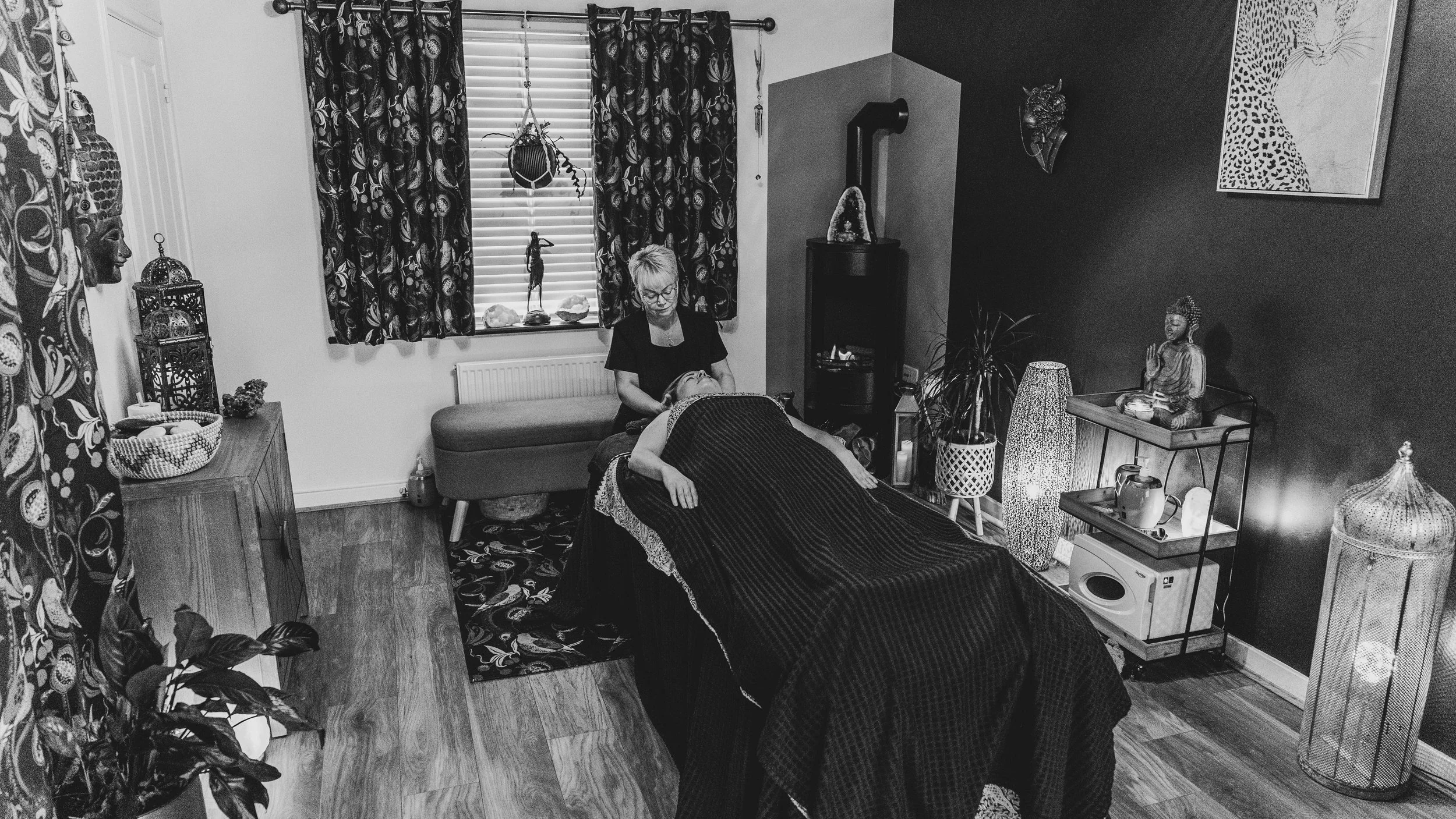 A woman is receiving a massage on a massage table while another woman operates the massage device. The room has patterned curtains, Asian-inspired decor, and various statues and plants.