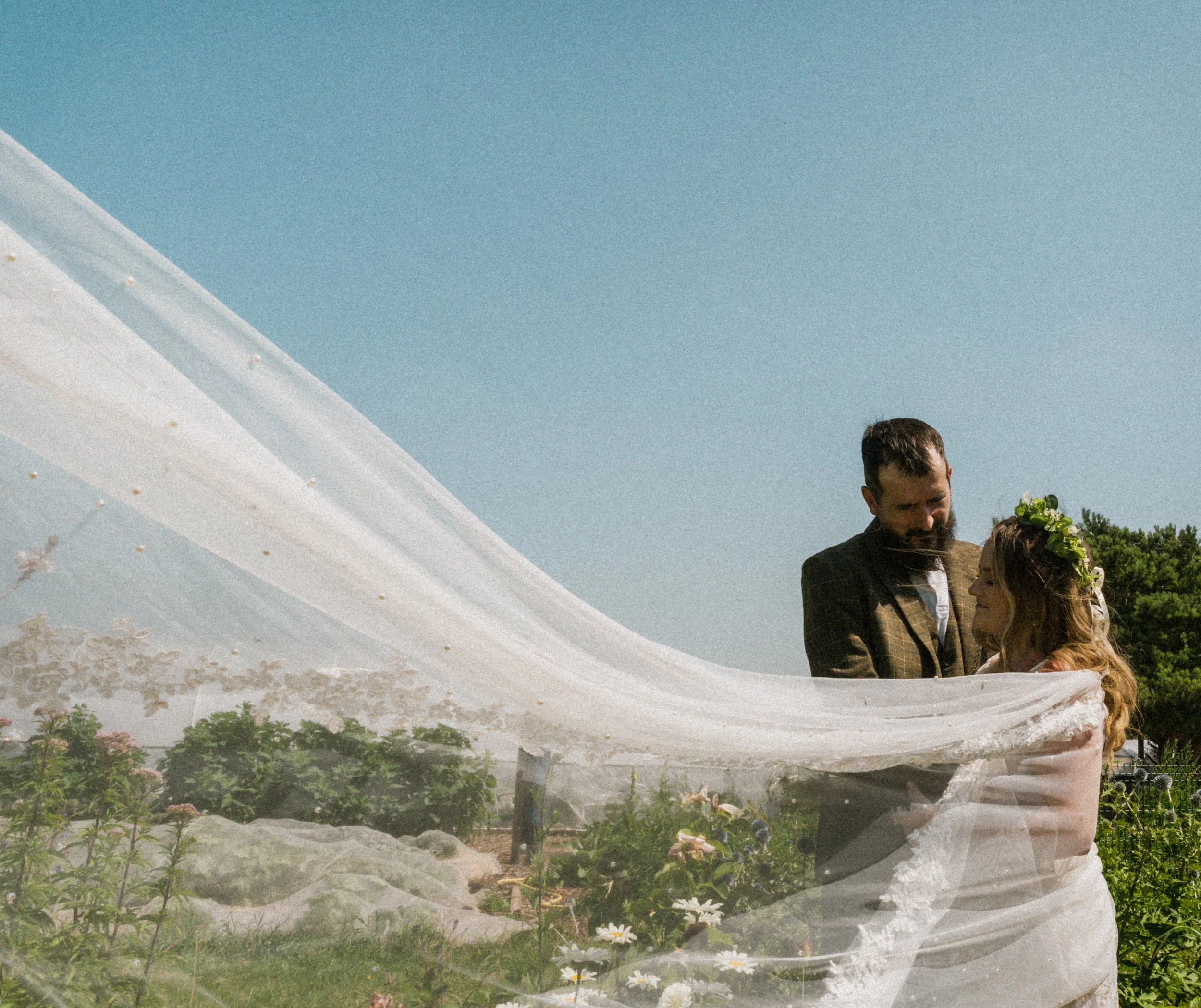 A couple with a bouquet of flowers standing under a white fabric canopy outdoors on a sunny day.