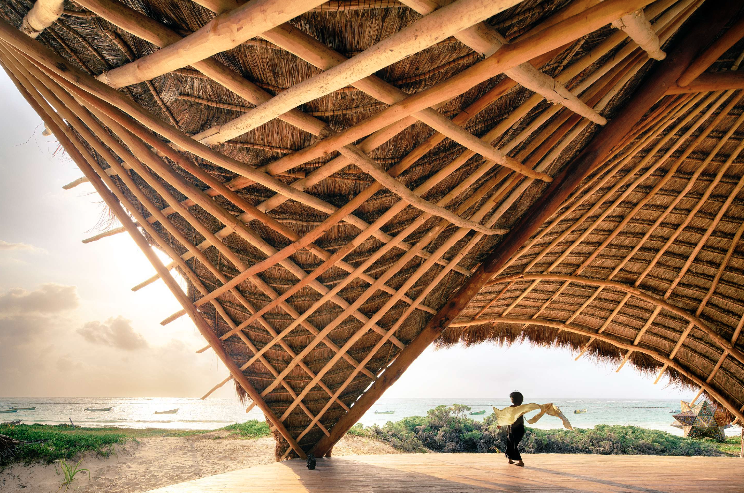 Person standing under a large thatched roof with a view of the beach and ocean in the background.