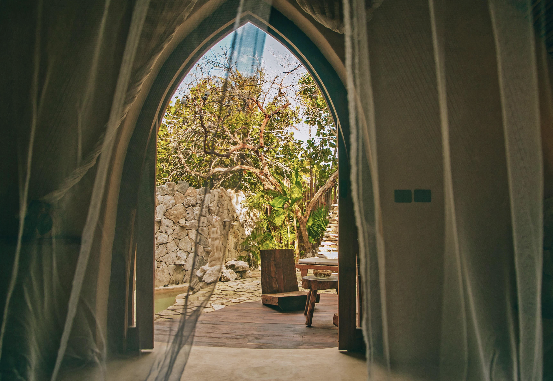 View through a doorway with sheer curtains onto a stone patio with outdoor furniture, surrounded by lush plants and trees, under a bright blue sky.