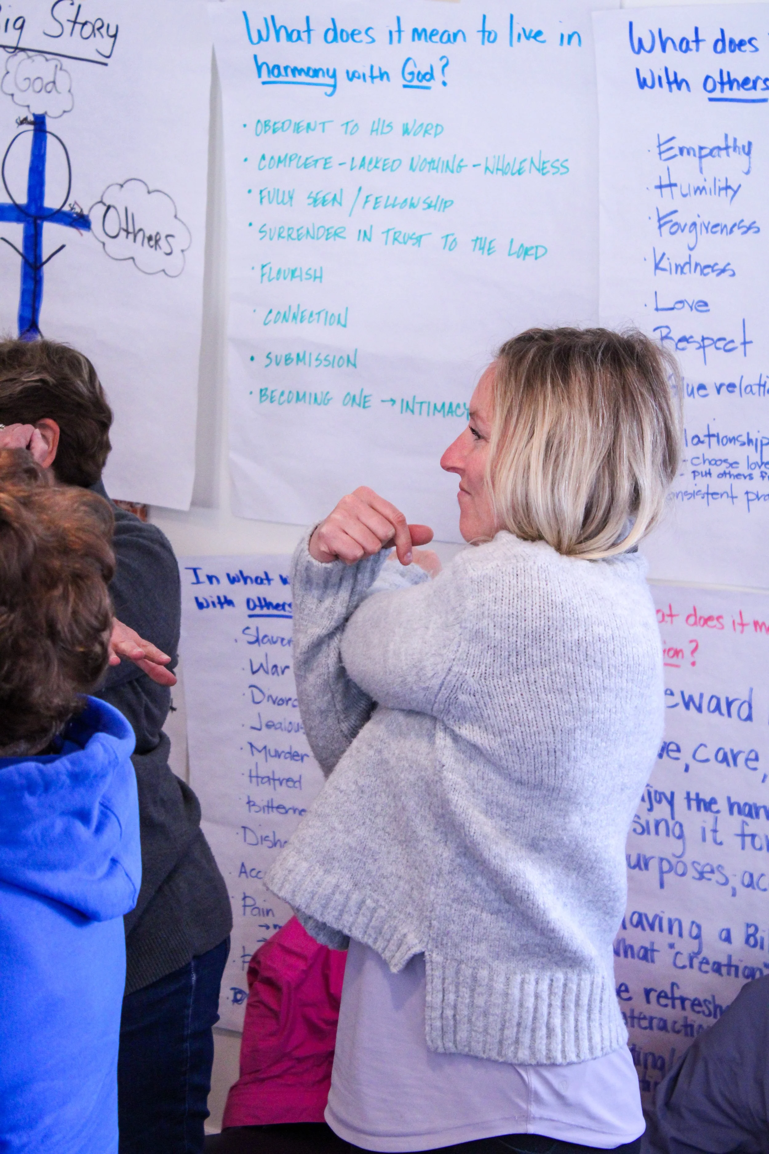 A woman with blonde hair and a gray sweater looks at a large poster with handwritten notes on a whiteboard. Several children with various jackets and scarves are also in the room, partially visible, gathered around the poster during a discussion or p