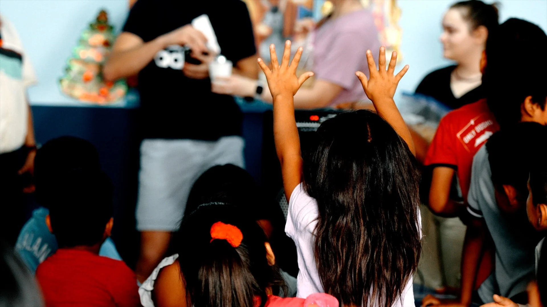 A young girl with long hair and a white shirt raises her hand during a classroom activity. Other children are seated around her, some with red or gray clothing, in a lively classroom setting.
