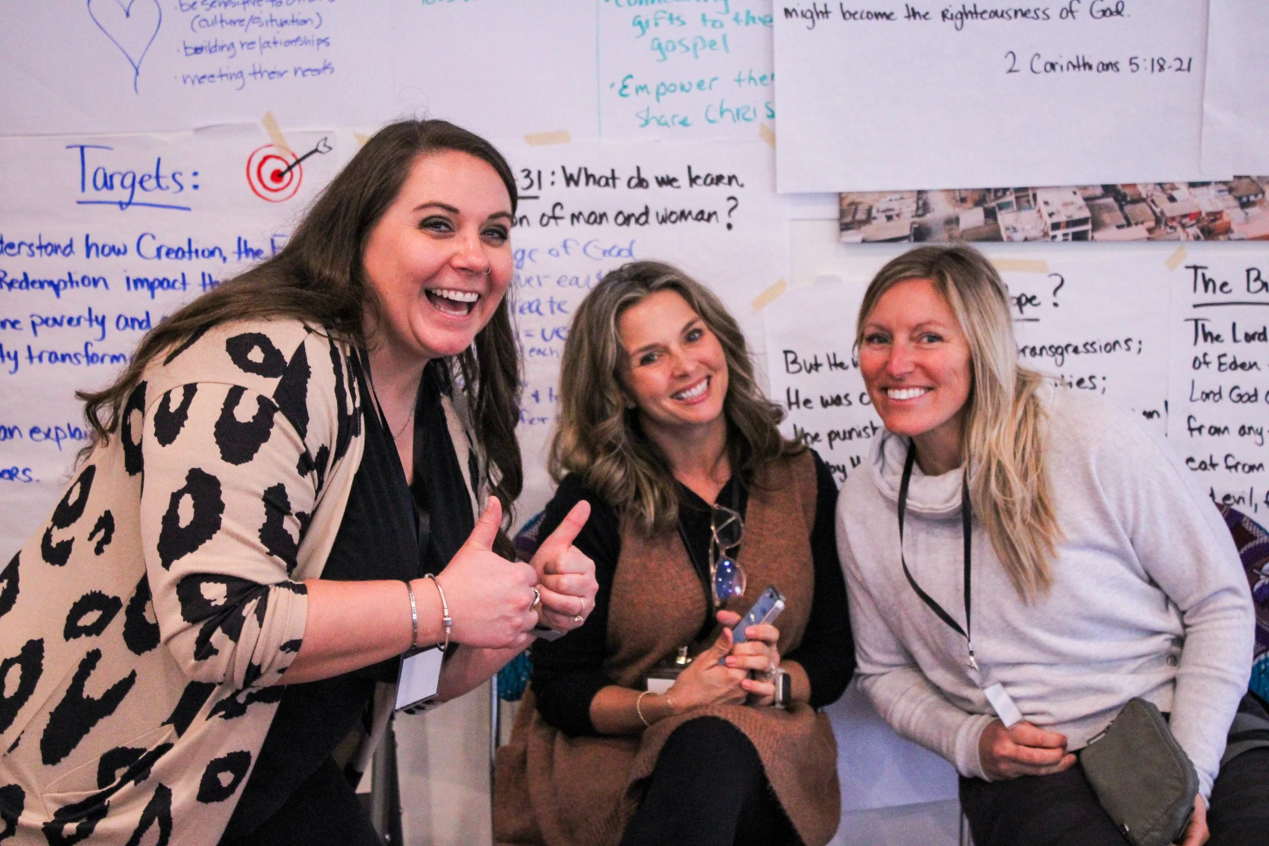 Three women smiling and giving thumbs up in front of a wall with handwritten notes and diagrams.