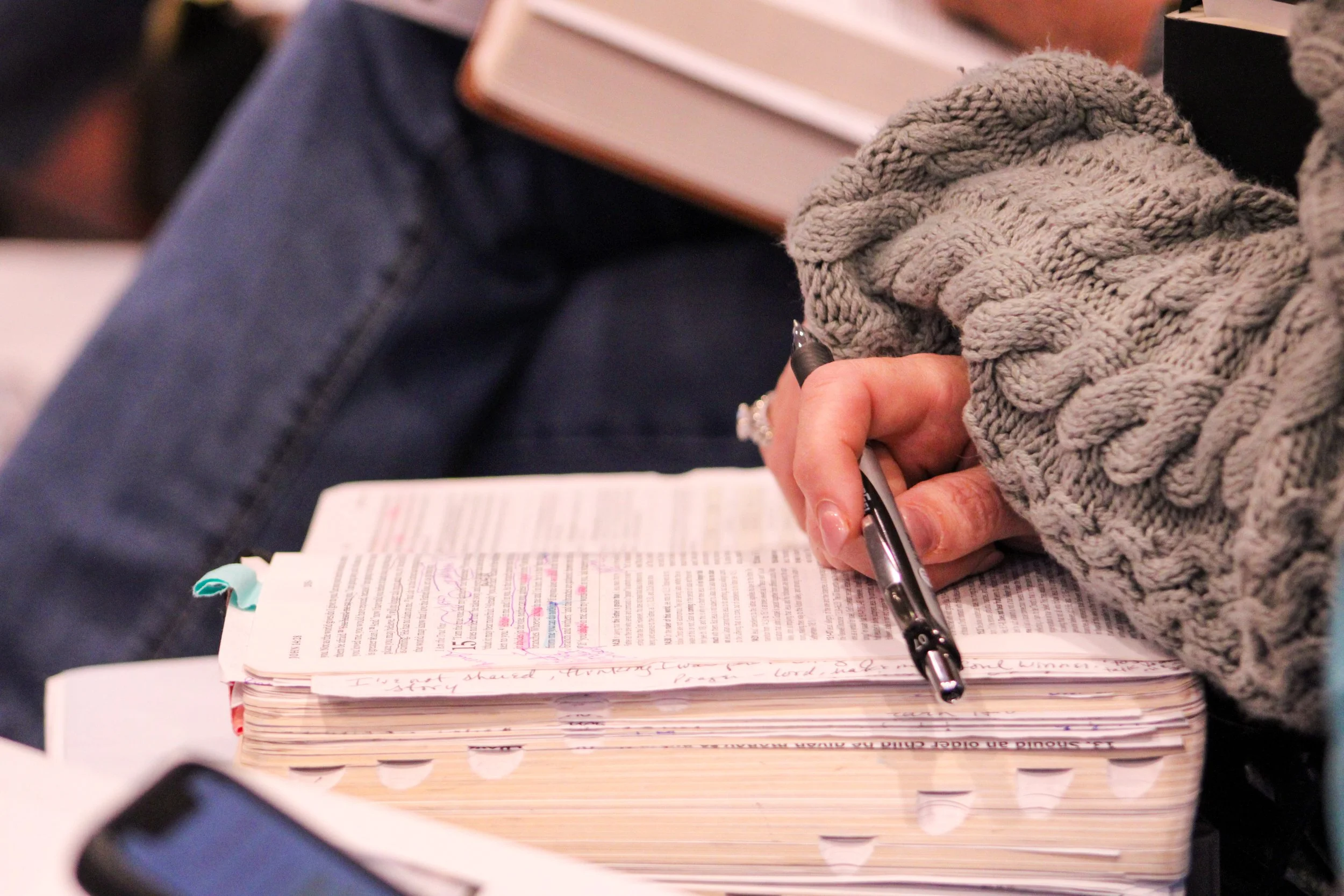 A person wearing a knitted gray sweater writing in a notebook with notes and highlighted text, sitting at a desk.