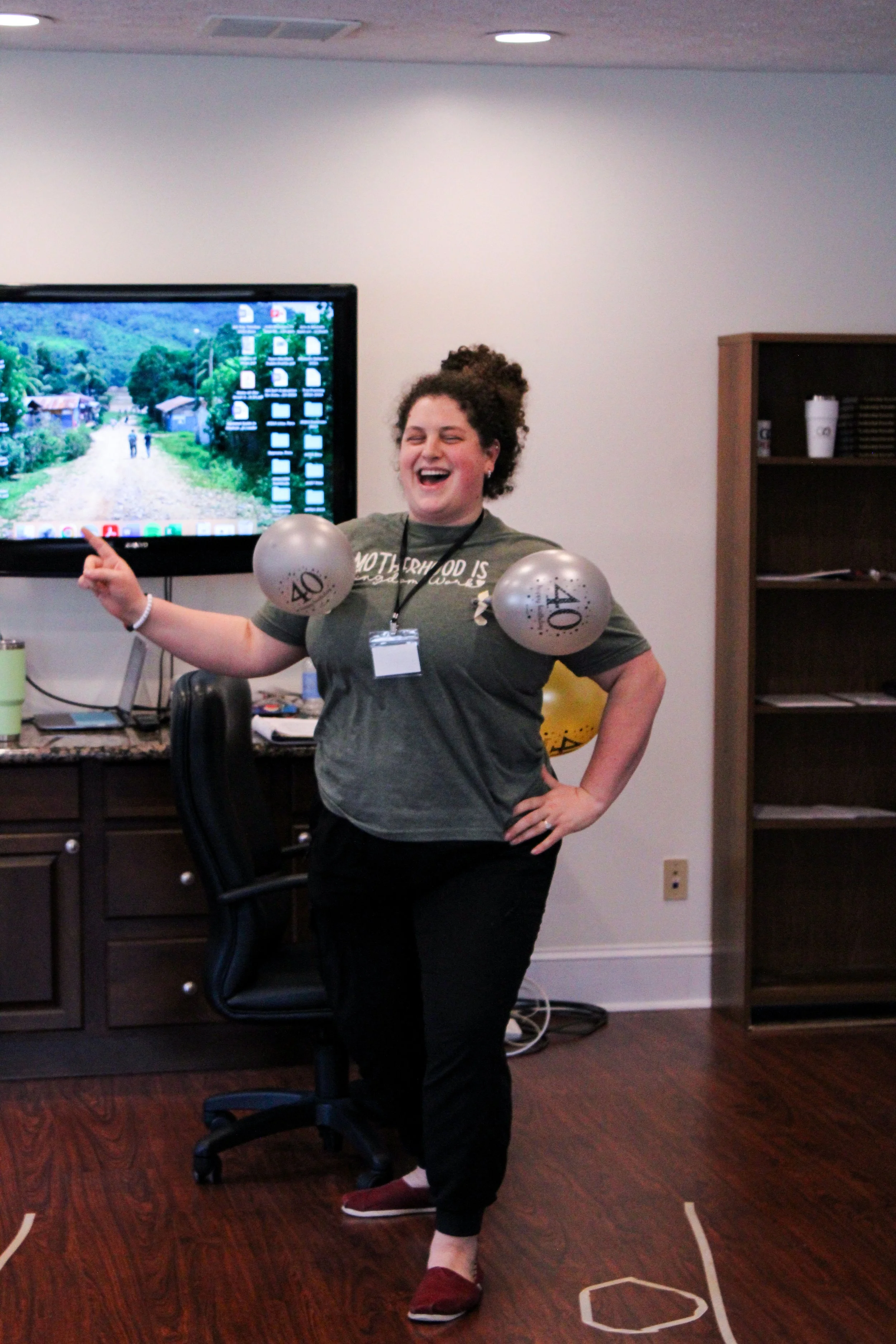 A woman with curly hair laughing and standing in an office, wearing a gray t-shirt, black pants, maroon shoes, and holding balloons with '40' on them, next to a large monitor and a wooden cabinet.
