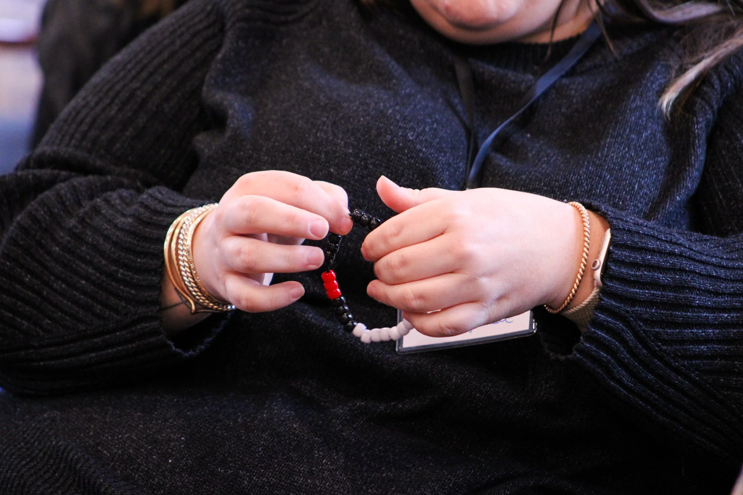 A person wearing a dark sweater holding a beaded bracelet in their hands, with gold and silver bracelets on their wrists.